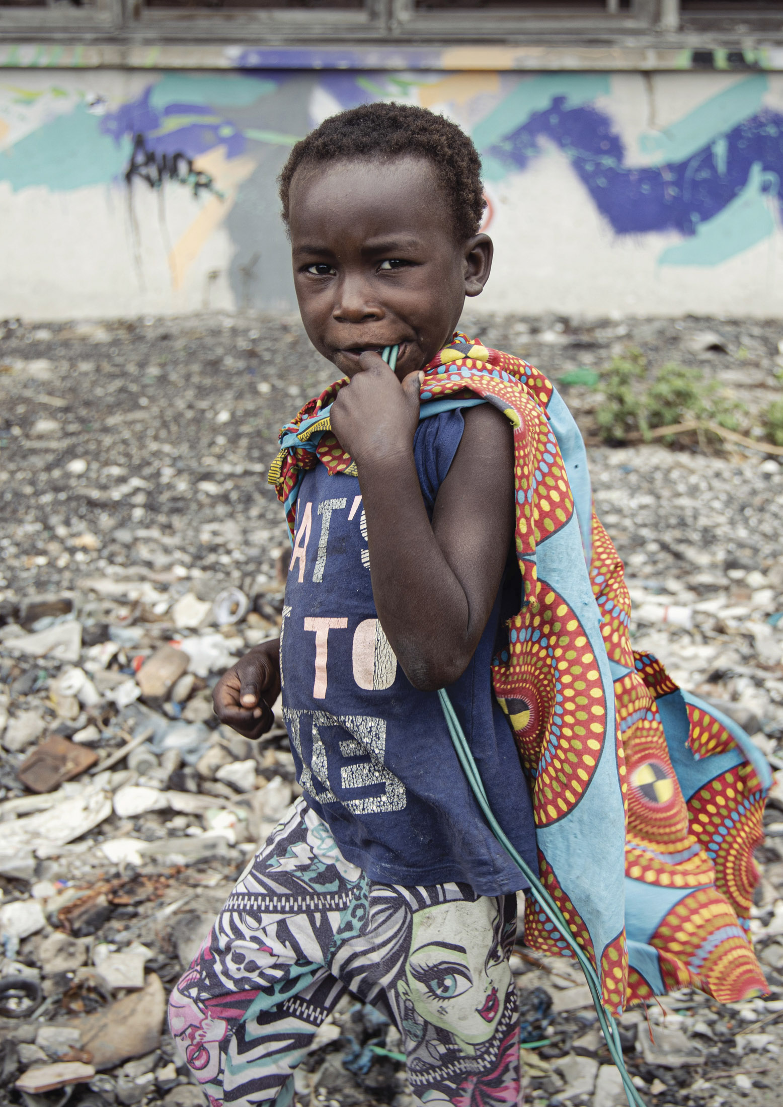 Ein kleiner Junge steht lächelnd auf einem mit Abfall übersäten Boden und trägt ein bunt bedrucktes Tuch über einem T-Shirt und einer gemusterten Hose. Im Hintergrund eine Graffitiwand. © Fotografie Tomas Rodriguez