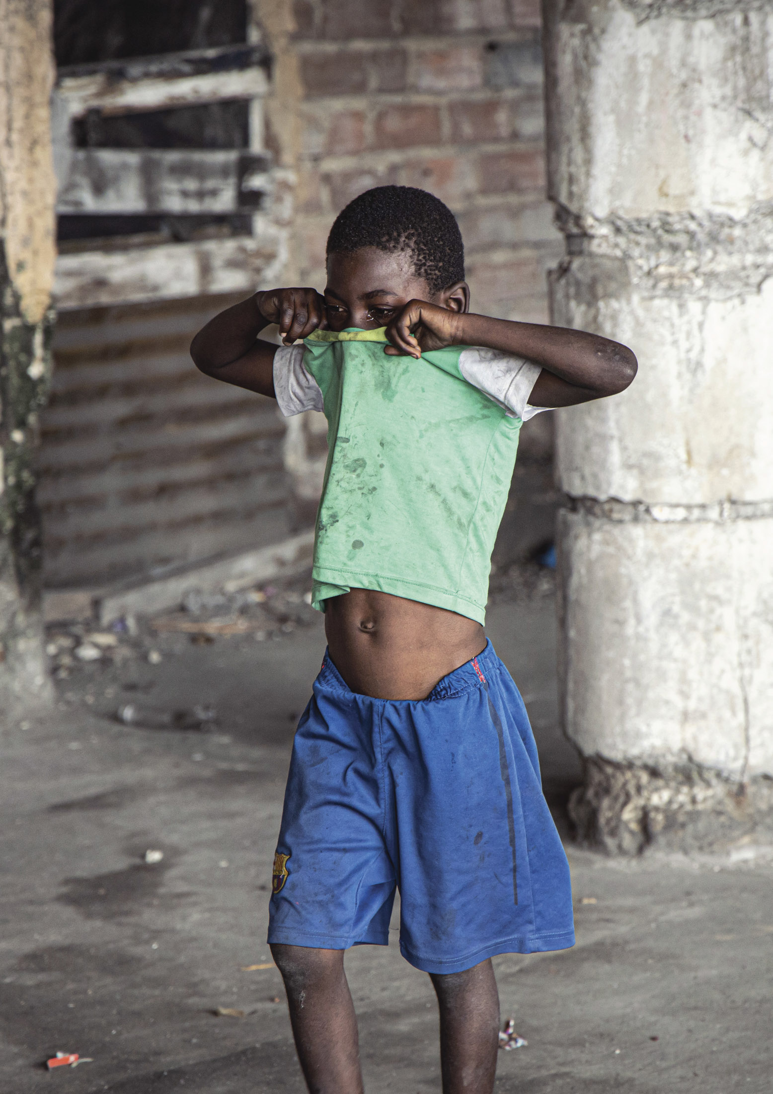 Ein Junge in einem grünen T-Shirt und blauen Shorts steht in einem heruntergekommenen Gebäude, hebt sein Hemd hoch, um sein Gesicht zu bedecken, und verschränkt die Arme. © Fotografie Tomas Rodriguez