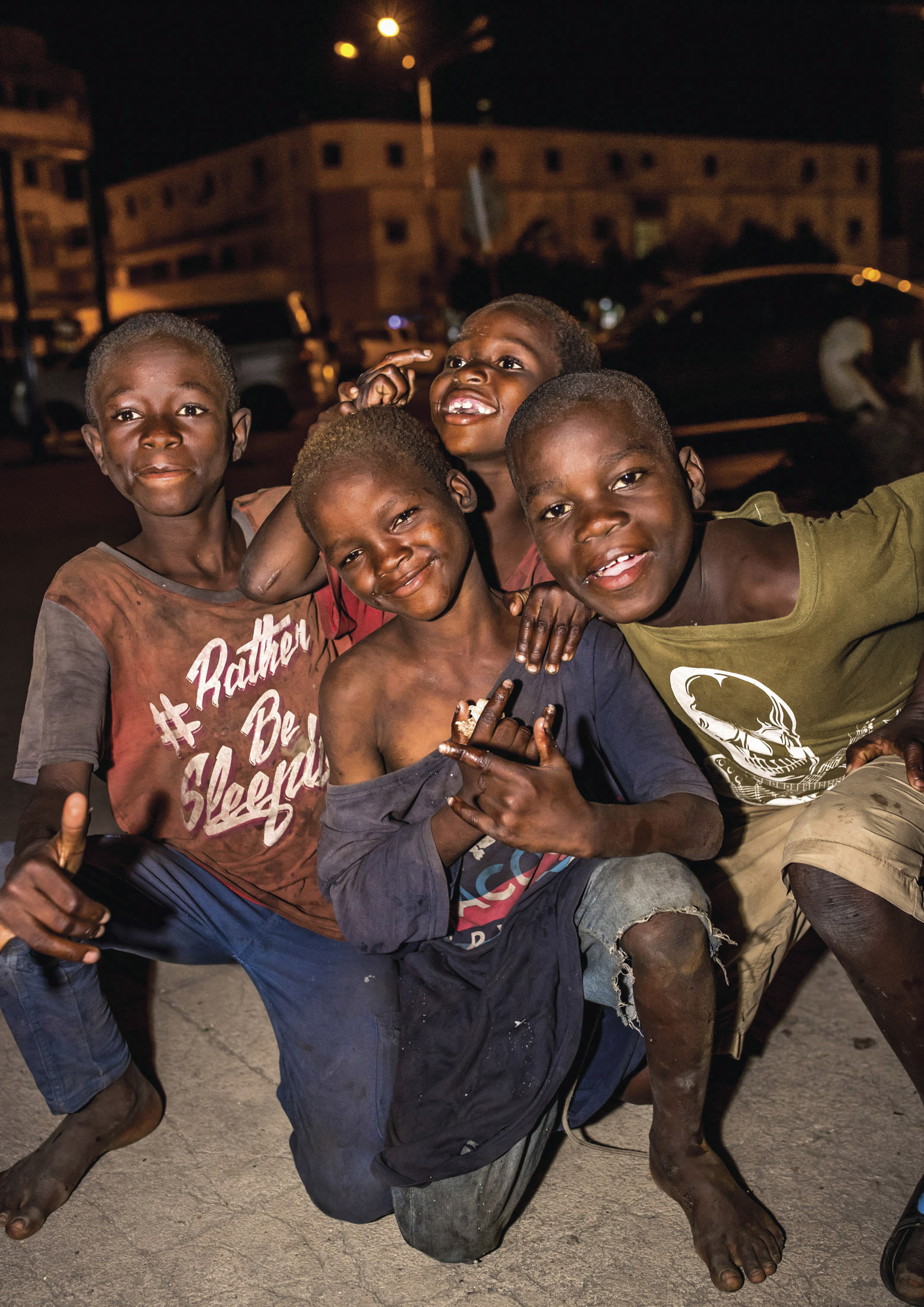 Vier fröhliche Jungen drängen sich nachts zusammen, lächeln und schauen in die Kamera. Einer hält Trommelstöcke, ein anderer zeigt seine Handflächen. Die Lichter der Stadt erhellen schwach den Hintergrund. © Fotografie Tomas Rodriguez