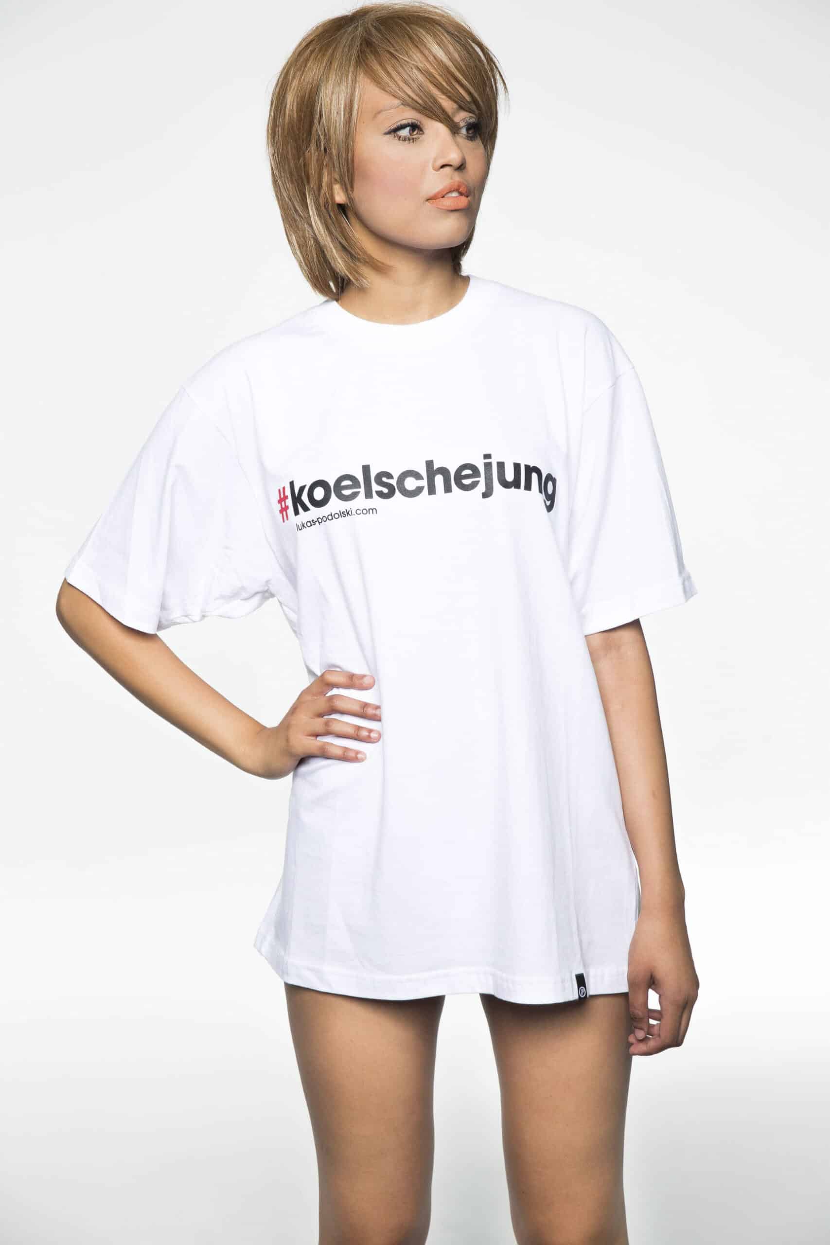 Eine junge Frau mit kurzen blonden Haaren, die ein übergroßes weißes T-Shirt mit der Aufschrift „Kölschejung“ trägt, steht vor einem schlichten weißen Hintergrund und blickt mit ernster Miene nach links. © Fotografie Tomas Rodriguez