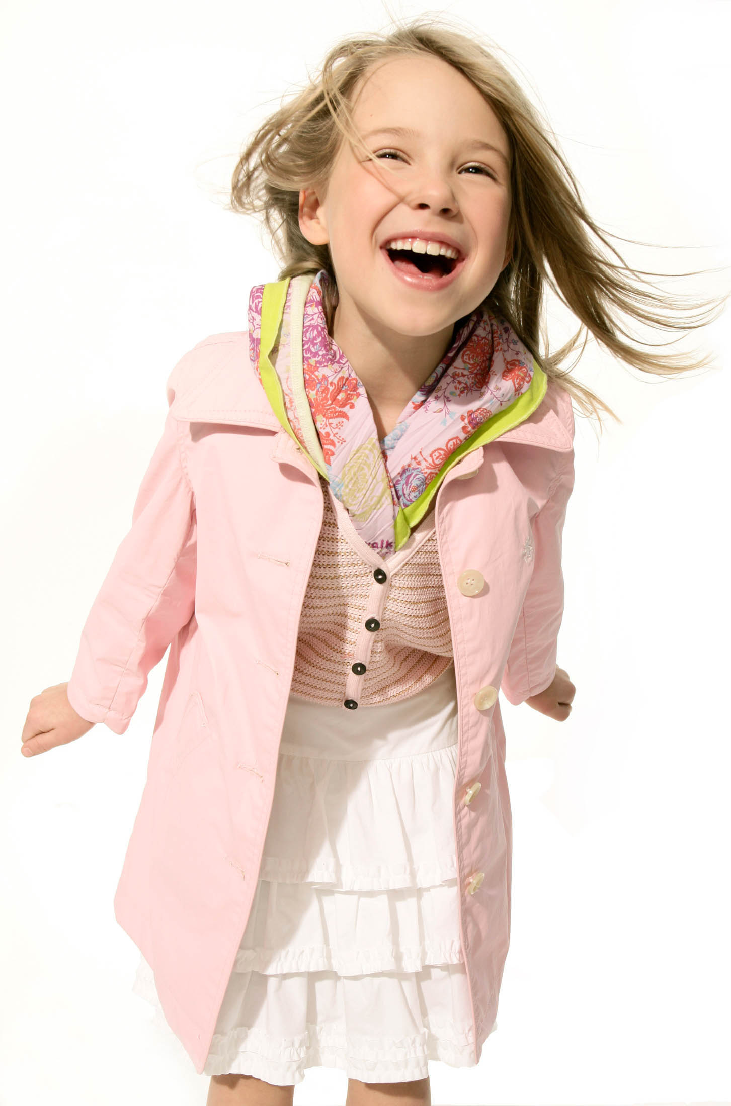 Ein fröhliches junges Mädchen mit wallendem blondem Haar, das einen hellrosa Mantel über einem weißen Kleid trägt, lacht, während sie vor einem weißen Hintergrund auf die Kamera zuläuft. © Fotografie Tomas Rodriguez