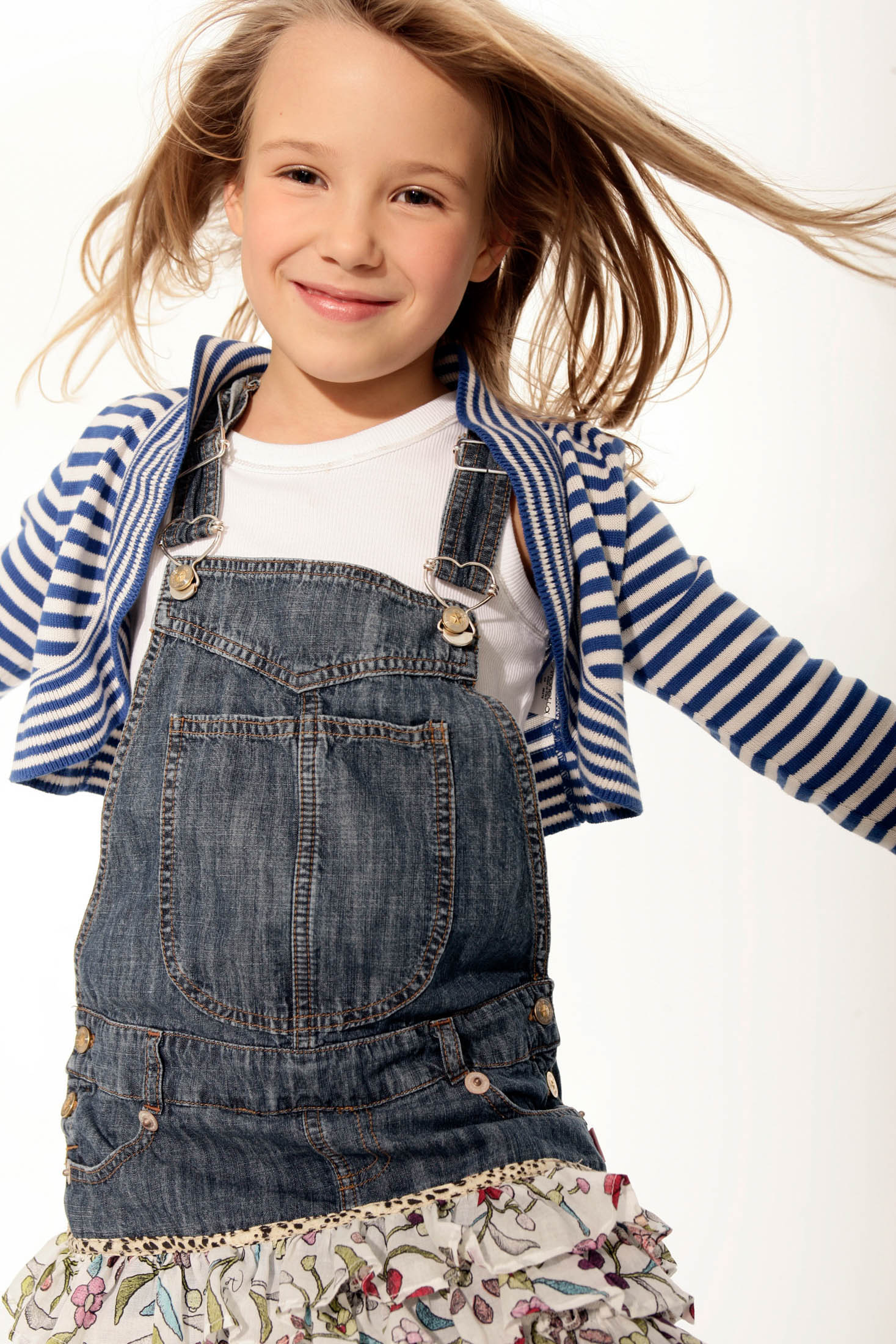 Ein fröhliches junges Mädchen mit wallendem Haar, einem gestreiften Hemd, einem Jeans-Overall und einem geblümten Rock, wirbelt fröhlich vor einem weißen Hintergrund herum. © Fotografie Tomas Rodriguez