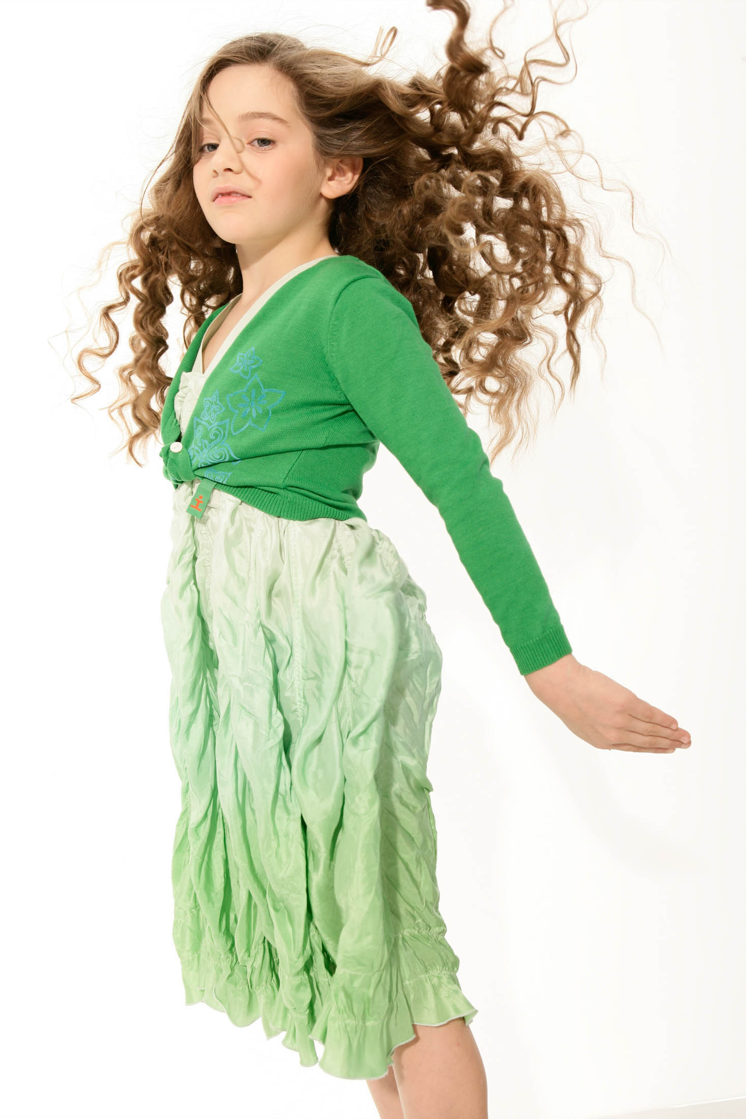 Ein junges Mädchen mit langen, lockigen Haaren in Bewegung, trägt eine grüne Strickjacke und ein hellgrünes, zerknittertes Kleid vor einem weißen Hintergrund. © Fotografie Tomas Rodriguez