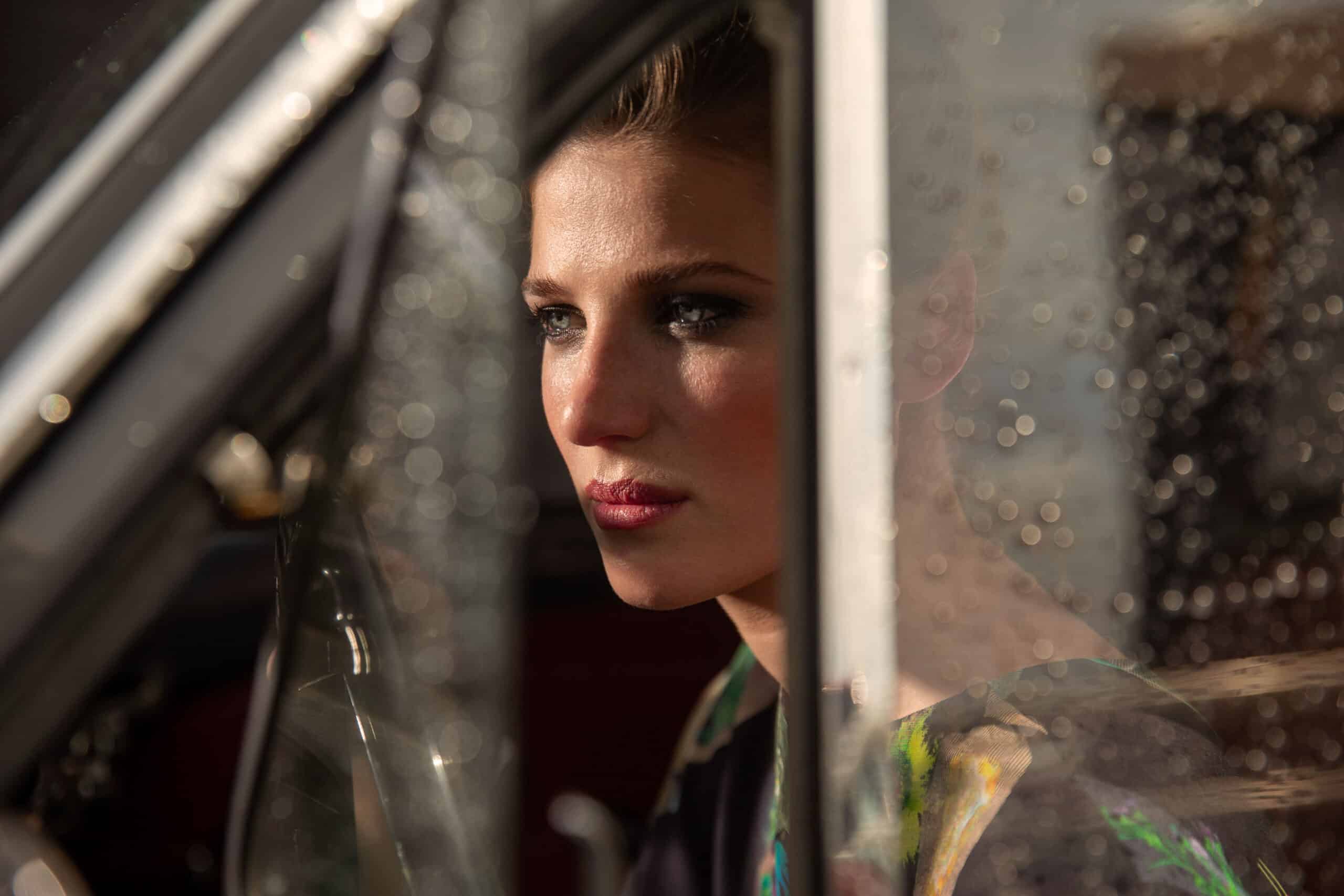 Eine Frau blickt nachdenklich aus einem Autofenster. Ihr Gesicht wird teilweise vom Sonnenlicht beleuchtet, was ihren klaren Teint und ihren nachdenklichen Ausdruck hervorhebt. © Fotografie Tomas Rodriguez