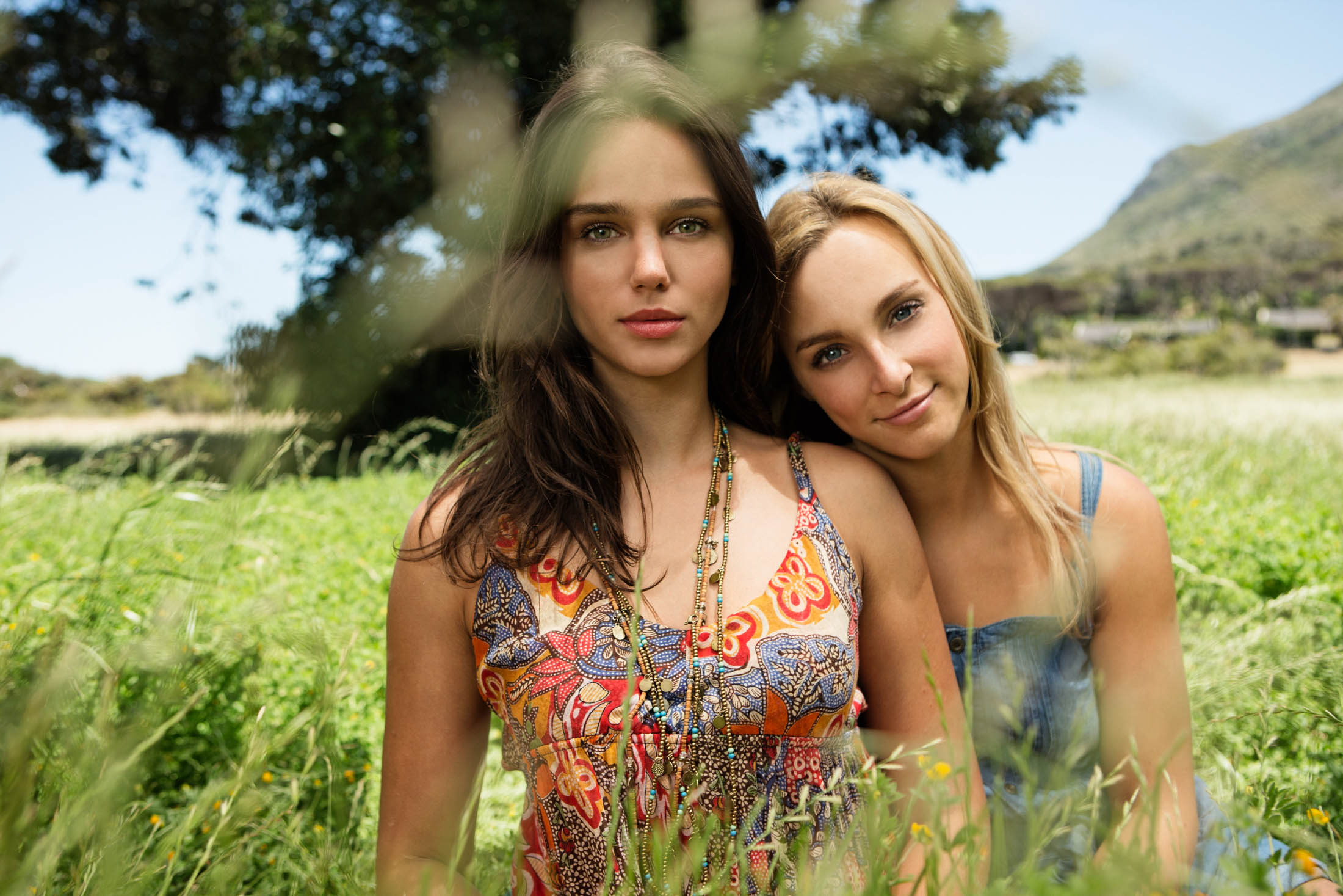 Zwei junge Frauen lächeln und posieren eng beieinander auf einer sonnigen Wildblumenwiese mit einem verschwommenen Berghintergrund. Eine trägt ein buntes Oberteil und die andere ein Jeansoberteil. © Fotografie Tomas Rodriguez