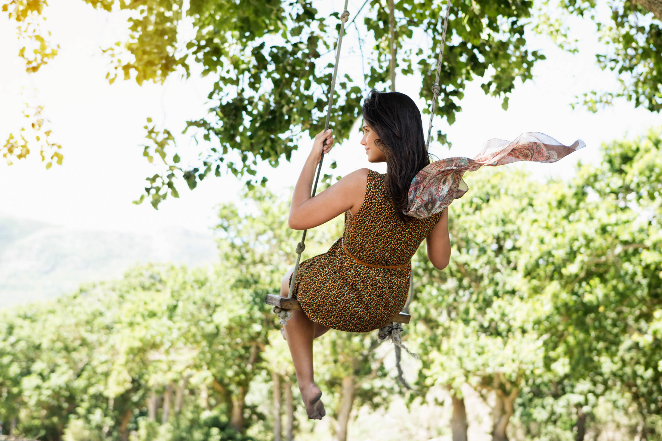 Eine Frau in einem gemusterten Kleid schaukelt auf einer Seilschaukel unter belaubten Bäumen, mit dem Rücken zur Kamera, während ihr Haar weht und die malerische Natur im Hintergrund zu sehen ist. © Fotografie Tomas Rodriguez