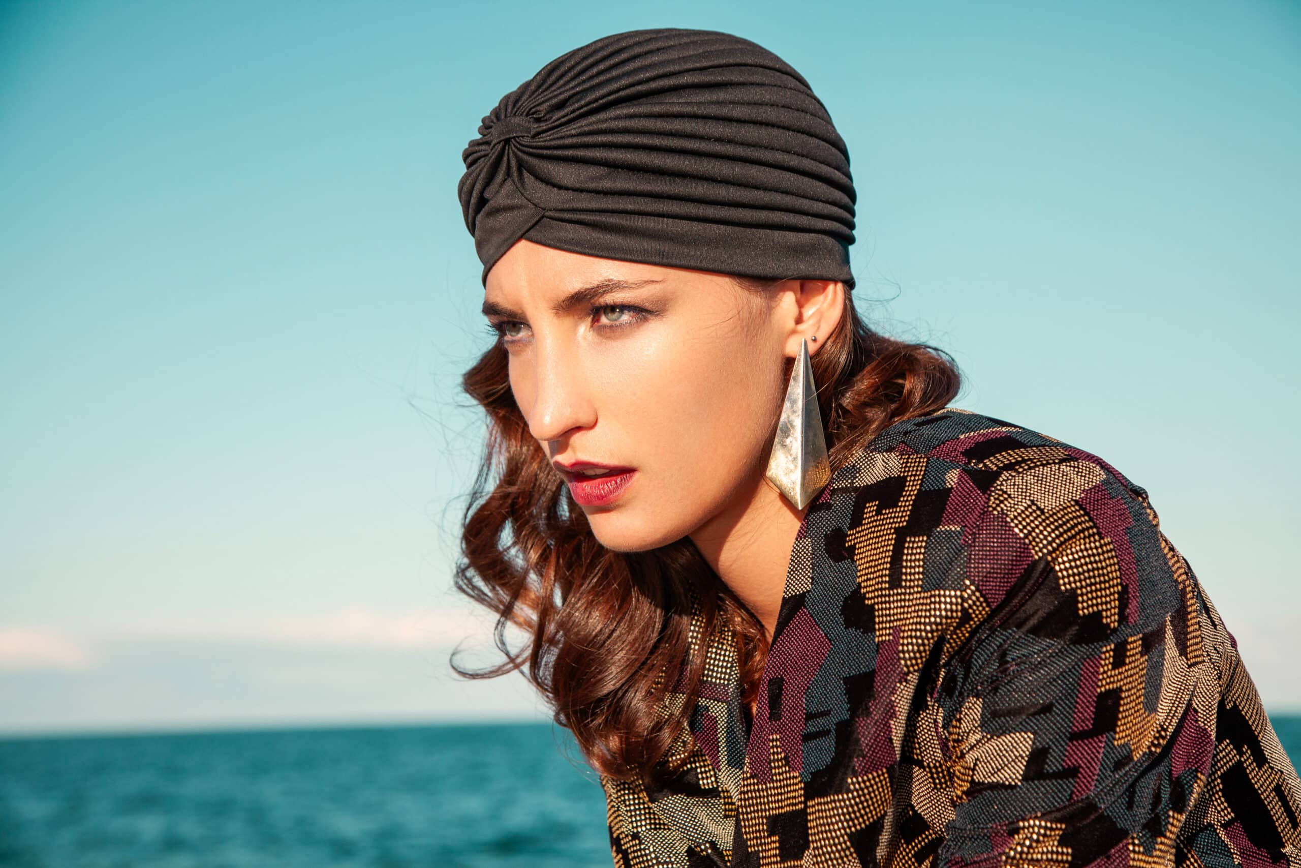 Eine Frau mit einem schicken Turban und einer gemusterten Jacke blickt nachdenklich vor dem Hintergrund eines klaren blauen Himmels und Ozeans. © Fotografie Tomas Rodriguez