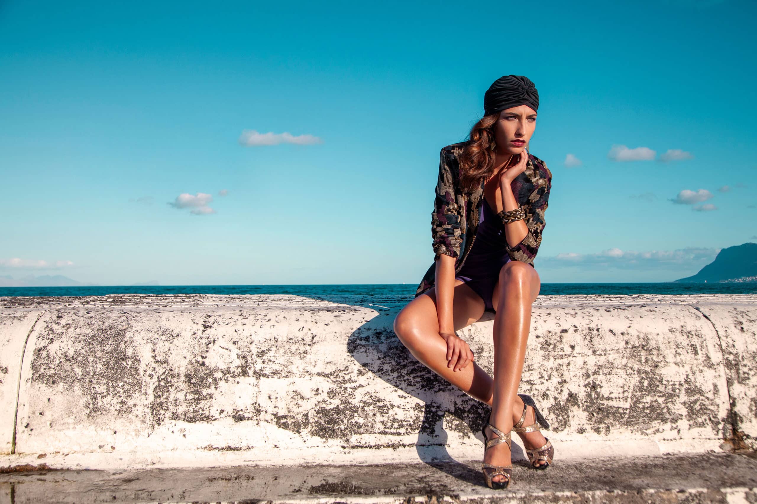 Eine Frau in einem modischen Outfit sitzt auf einer verwitterten weißen Wand, hinter ihr das Meer und ein klarer blauer Himmel. Sie trägt ein Kopftuch und High Heels und blickt in die Ferne. © Fotografie Tomas Rodriguez