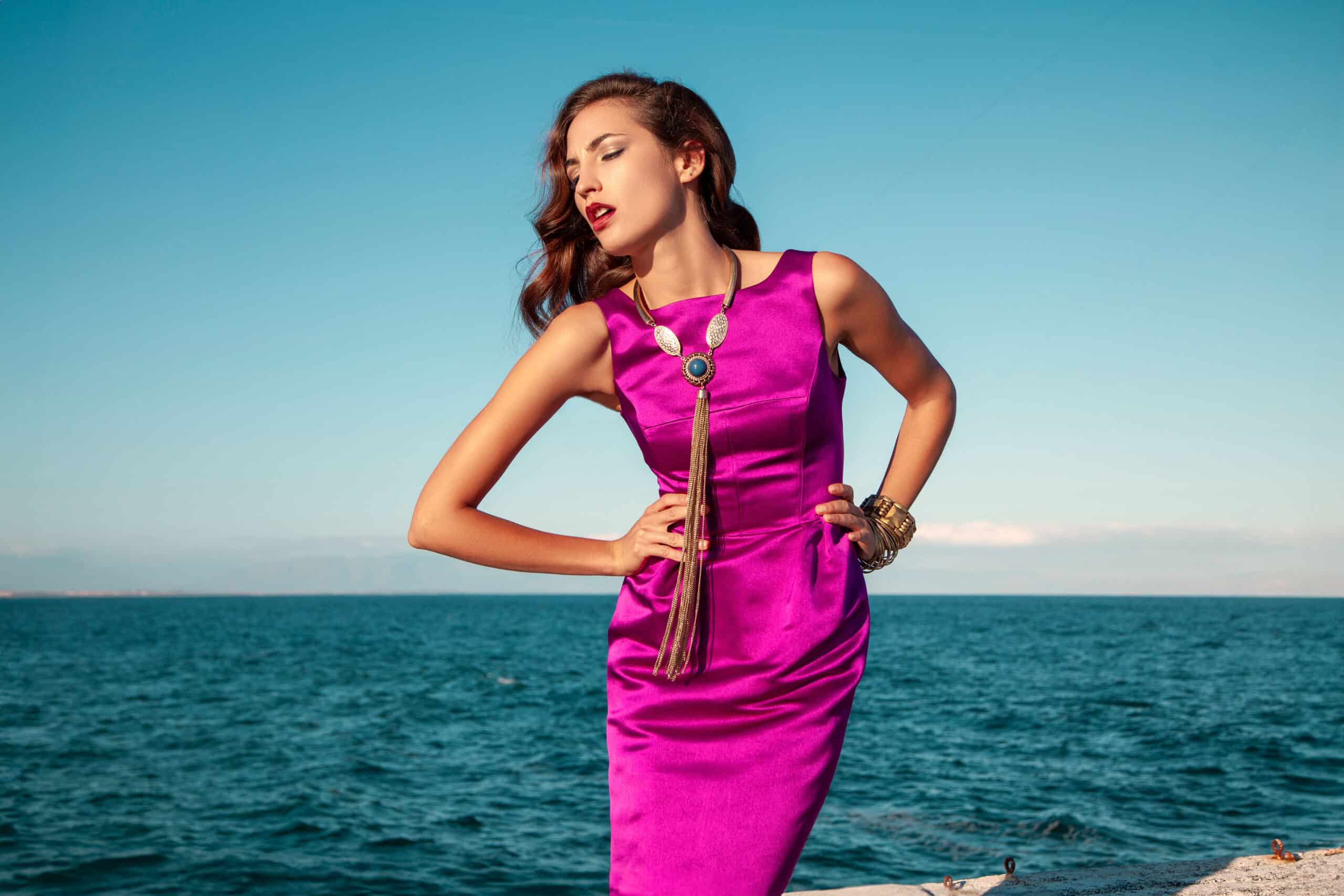 Eine Frau in einem leuchtend rosa Kleid steht elegant am Meer, mit einem klaren blauen Himmel im Hintergrund, ihr Haar ist zur Seite frisiert. Sie posiert selbstbewusst, die Hand auf der Hüfte. © Fotografie Tomas Rodriguez