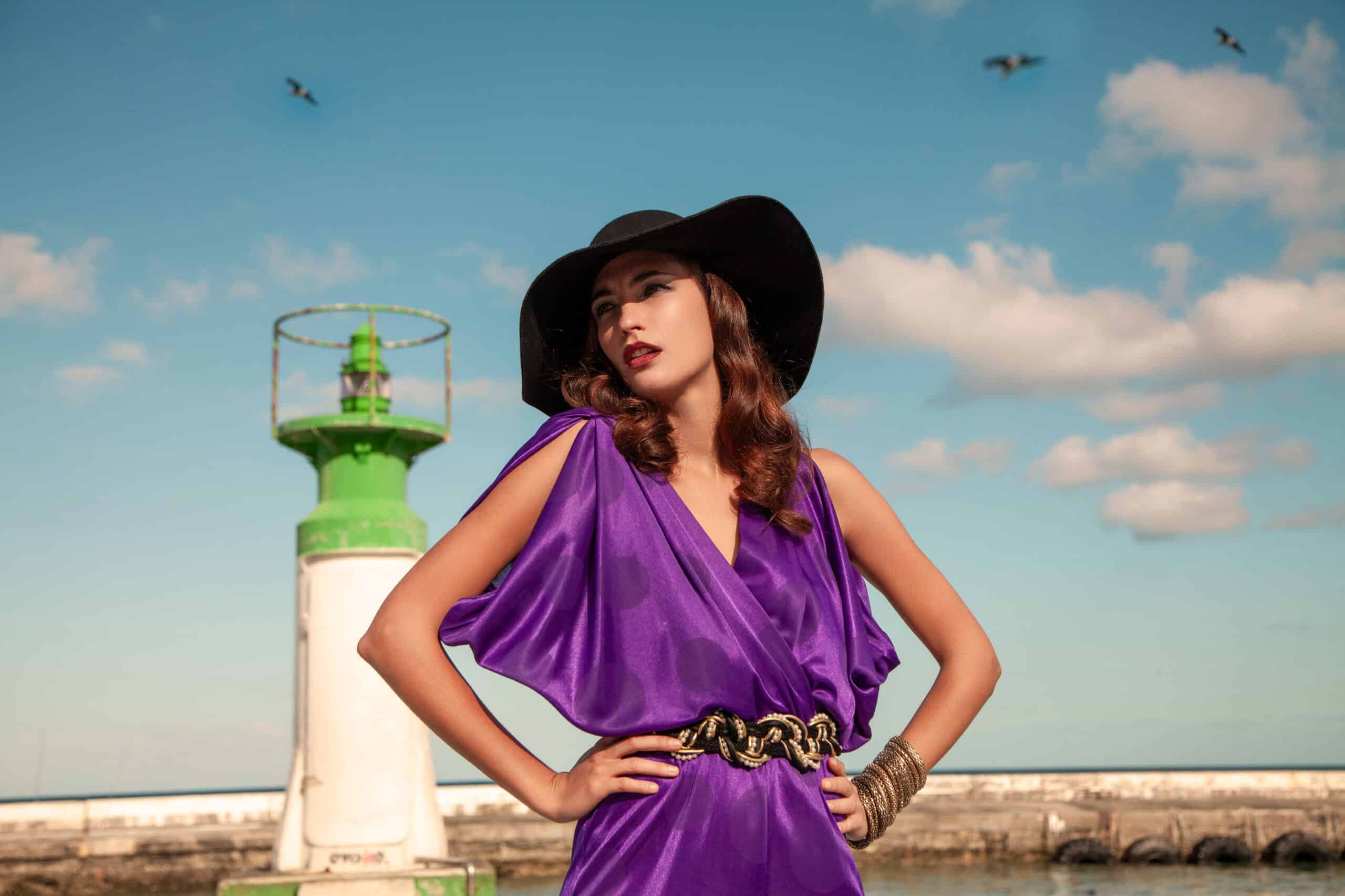 Eine Frau in einem lila Kleid und einem schwarzen Hut steht selbstbewusst neben einem grünen Leuchtturm, im Hintergrund ein klarer blauer Himmel und fliegende Vögel. © Fotografie Tomas Rodriguez