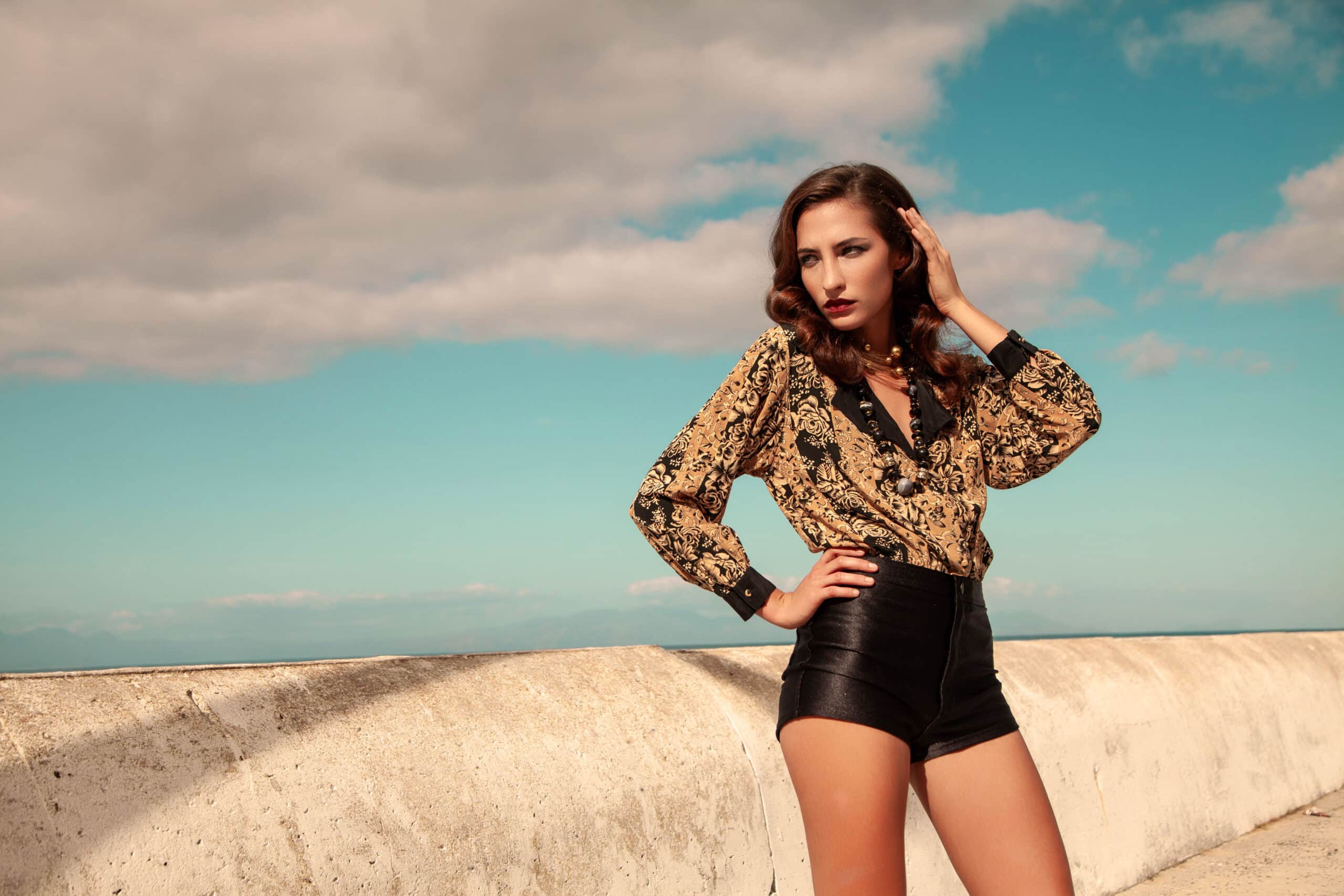 Ein Model posiert selbstbewusst auf einem Dach, gekleidet in eine gemusterte gold-schwarze Bluse und schwarze Shorts, mit einem dramatischen Wolkenhimmel und Horizont im Hintergrund. © Fotografie Tomas Rodriguez