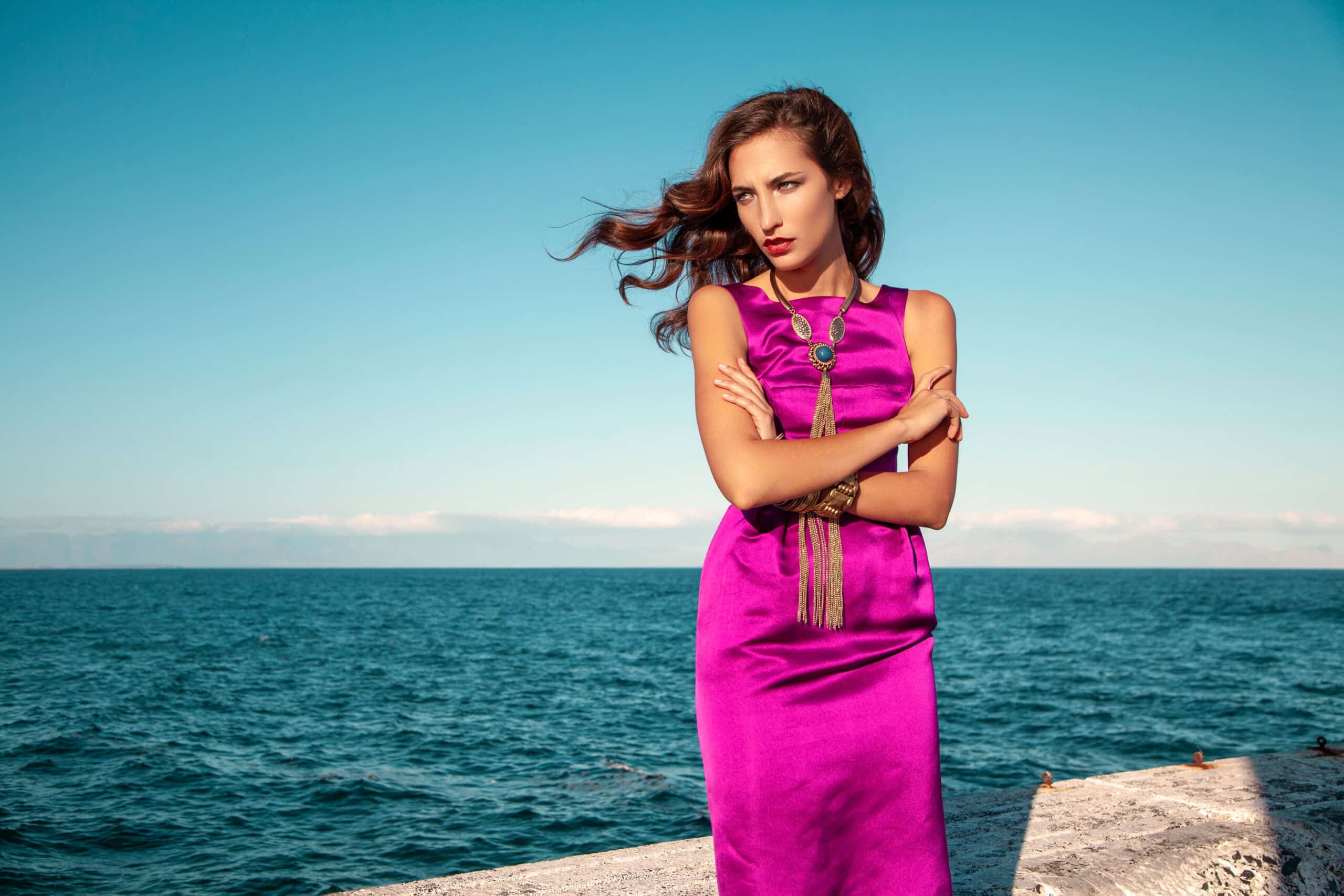 Eine Frau in einem leuchtend lila Kleid steht mit verschränkten Armen, vom Wind zerzaustem Haar und strenger Miene vor der Kulisse eines strahlend blauen Himmels und Meeres an einer Strandpromenade. © Fotografie Tomas Rodriguez