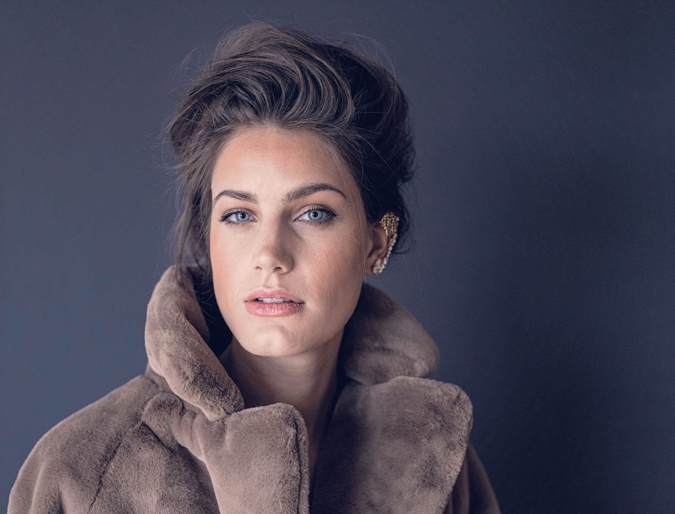 Ein Porträt einer Frau mit auffallend blauen Augen, die einen weichen braunen Mantel mit hohem Kragen trägt und ihr Haar elegant frisiert ist, vor einem grauen Hintergrund. © Fotografie Tomas Rodriguez