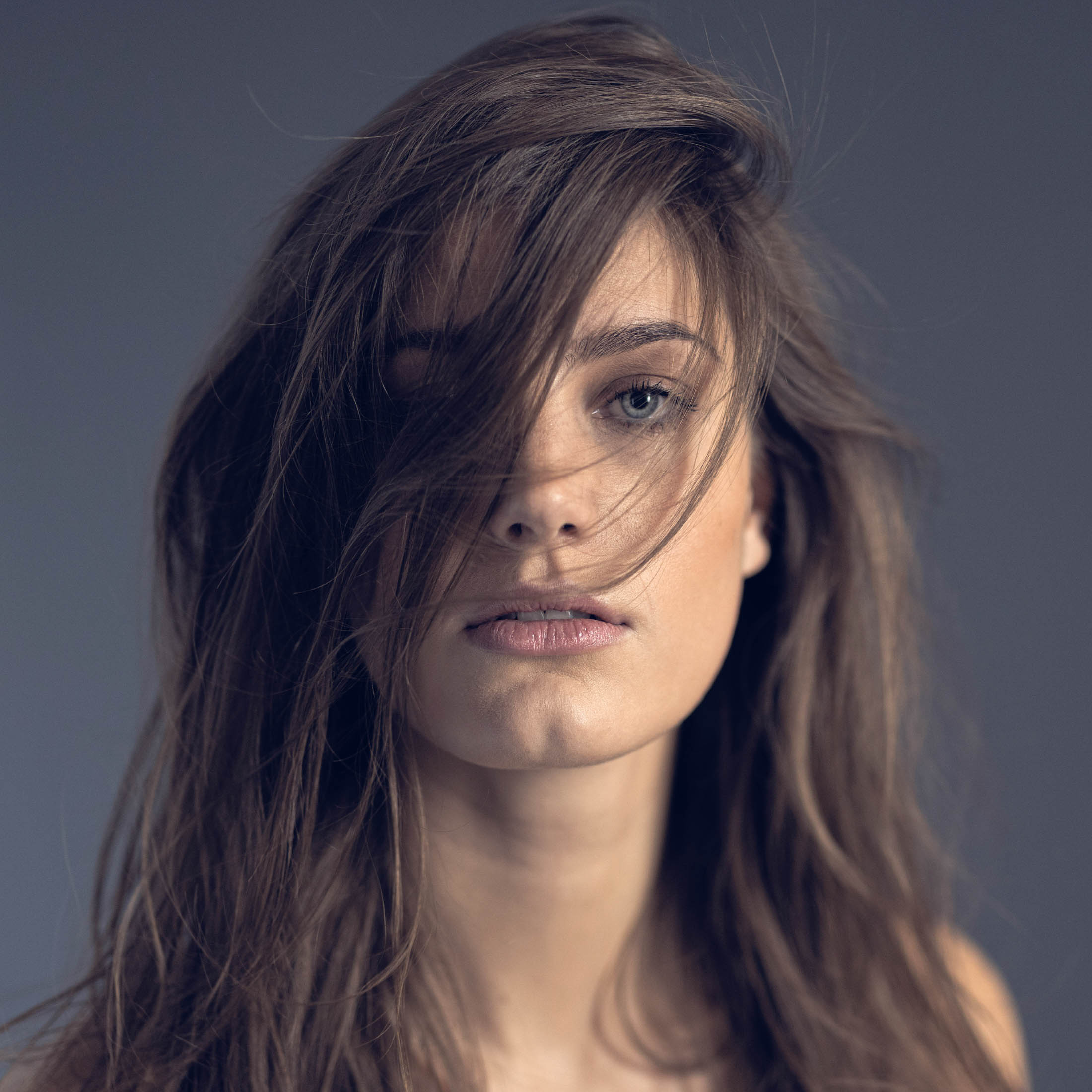 Porträt einer Frau mit zerzaustem braunem Haar, das ihr Gesicht teilweise bedeckt, und vor einem neutralen grauen Hintergrund intensiv in die Kamera blickt. © Fotografie Tomas Rodriguez