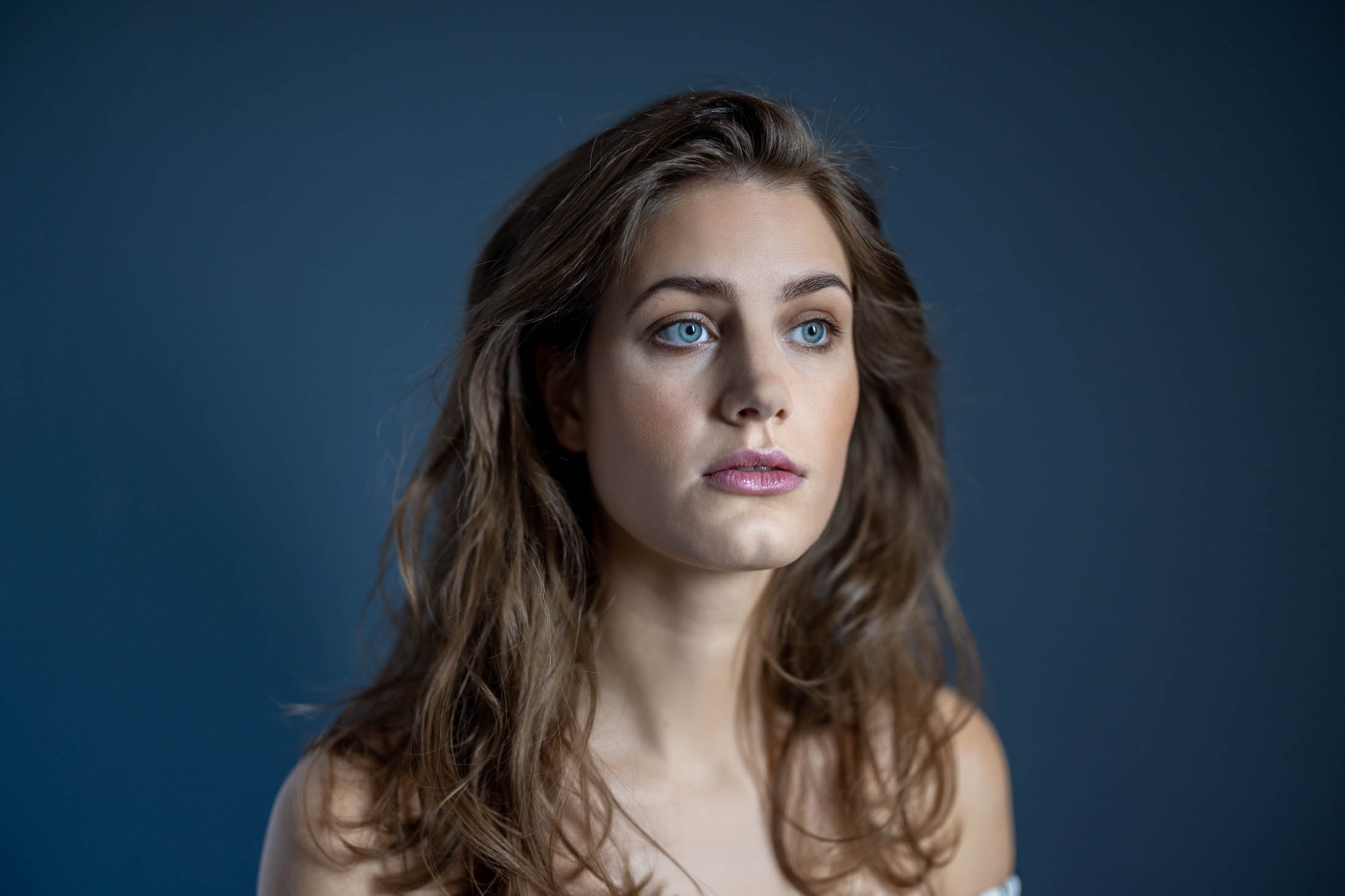 Ein Porträt einer jungen Frau mit langen welligen Haaren und dezentem Make-up, die vor einem blauen Hintergrund nachdenklich zur Seite blickt. © Fotografie Tomas Rodriguez