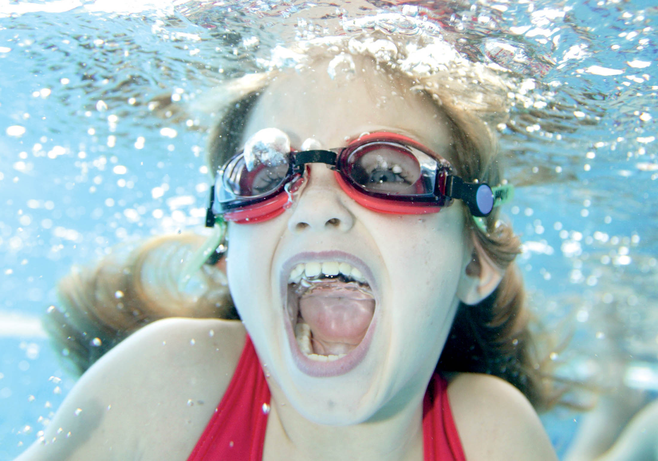 Ein junges Mädchen mit roter Schwimmbrille schwimmt unter Wasser, streckt spielerisch die Zunge heraus und hat dabei Blasen im Gesicht. © Fotografie Tomas Rodriguez