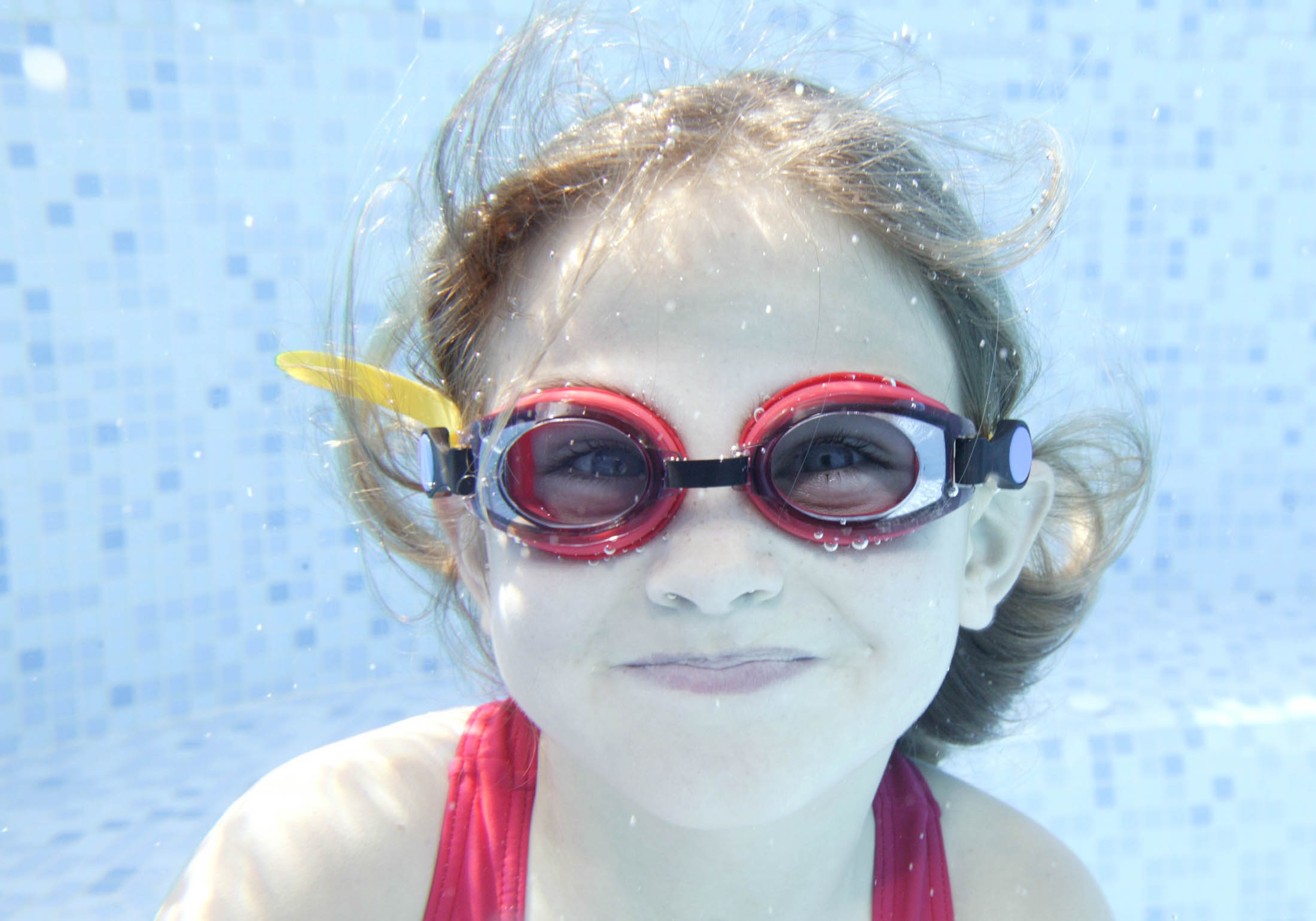 Ein junges Mädchen mit einer roten Schwimmbrille und einem Lächeln schwimmt unter Wasser in einem klaren blauen Becken mit sichtbaren Mosaikfliesen im Hintergrund. © Fotografie Tomas Rodriguez