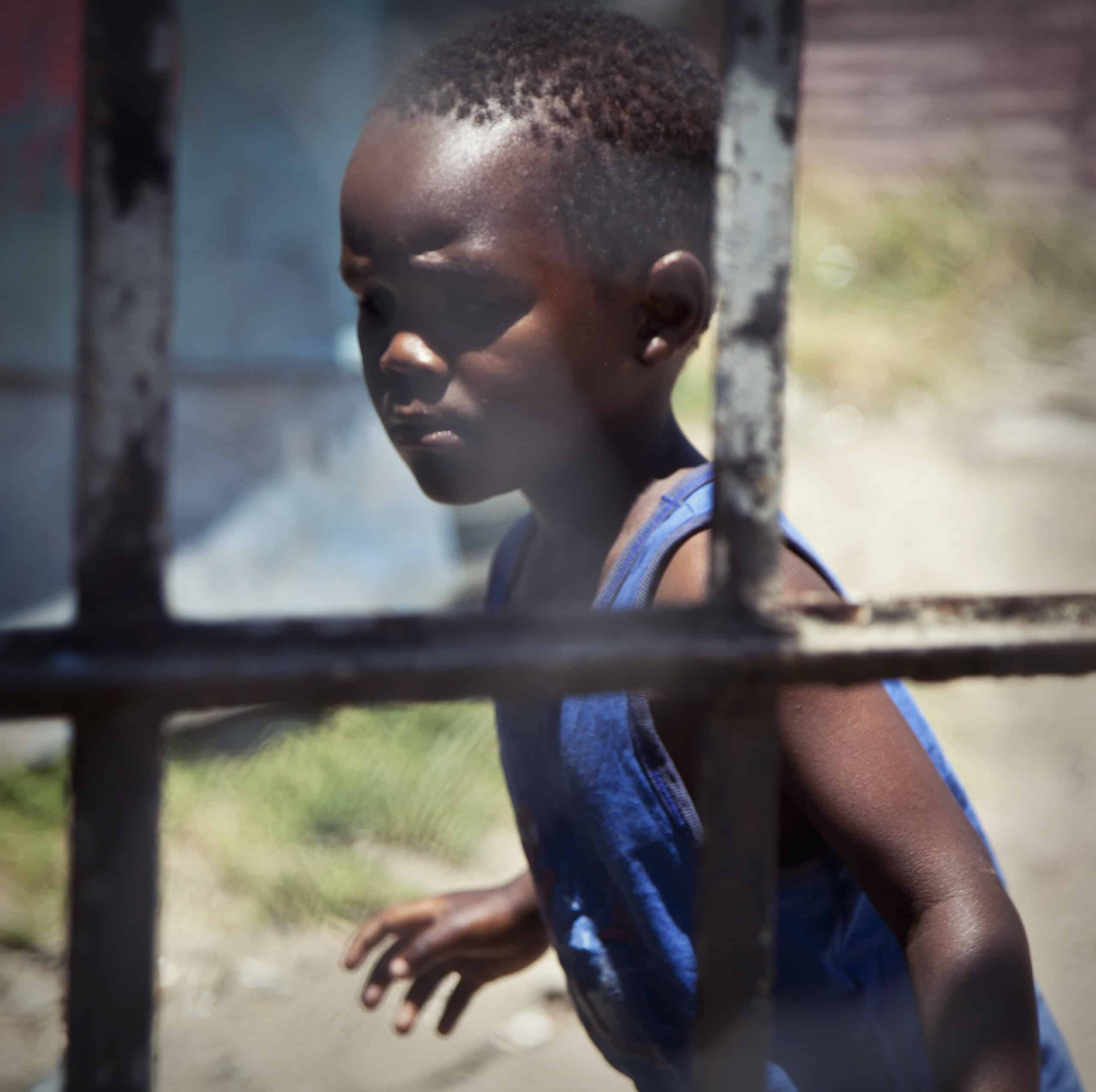 Ein kleiner Junge in einem blauen Tanktop steht hinter einem Metallgitter und blickt nachdenklich zur Seite. Das Bild ist leicht unscharf, was der Szene eine verträumte Qualität verleiht. © Fotografie Tomas Rodriguez