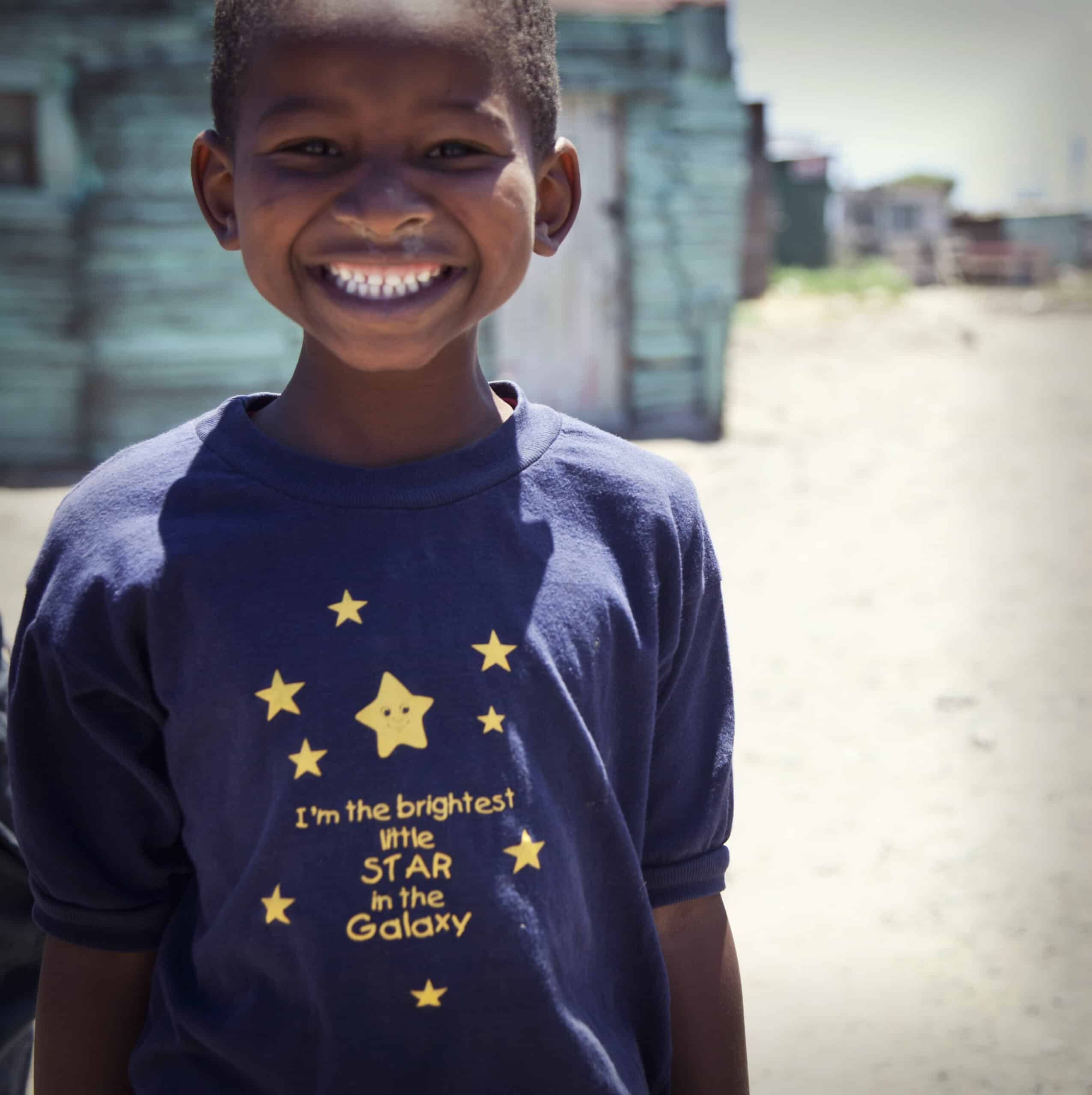 Ein fröhlicher kleiner Junge mit einem strahlenden Lächeln und einem blauen T-Shirt mit der Aufschrift „Ich bin der hellste kleine Stern in der Galaxie“ steht im Freien, im Hintergrund sind verschwommene Gebäude zu sehen. © Fotografie Tomas Rodriguez