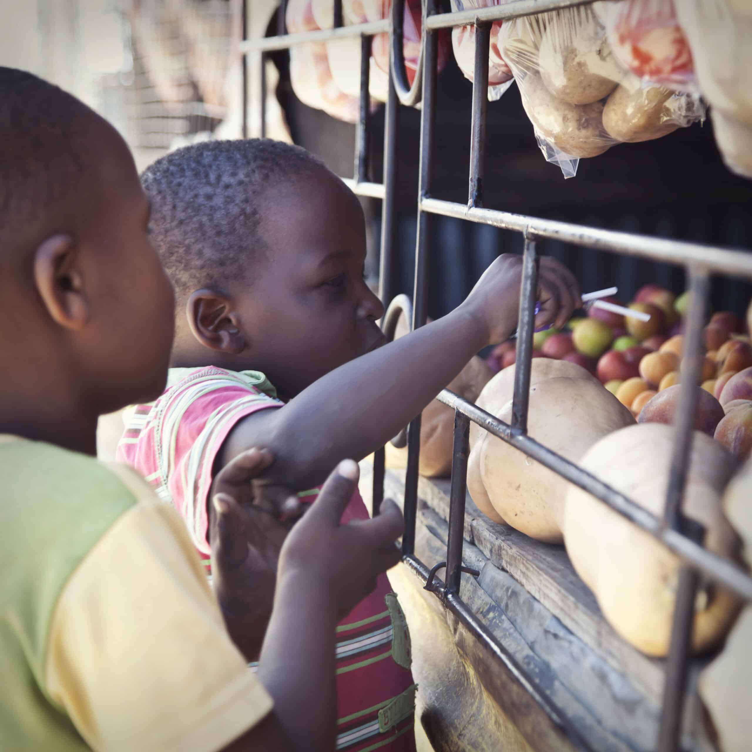 Zwei Kinder, eines greift nach Obst, stehen vor einem Straßenstand voller frischer Produkte und abgepackter Waren. © Fotografie Tomas Rodriguez