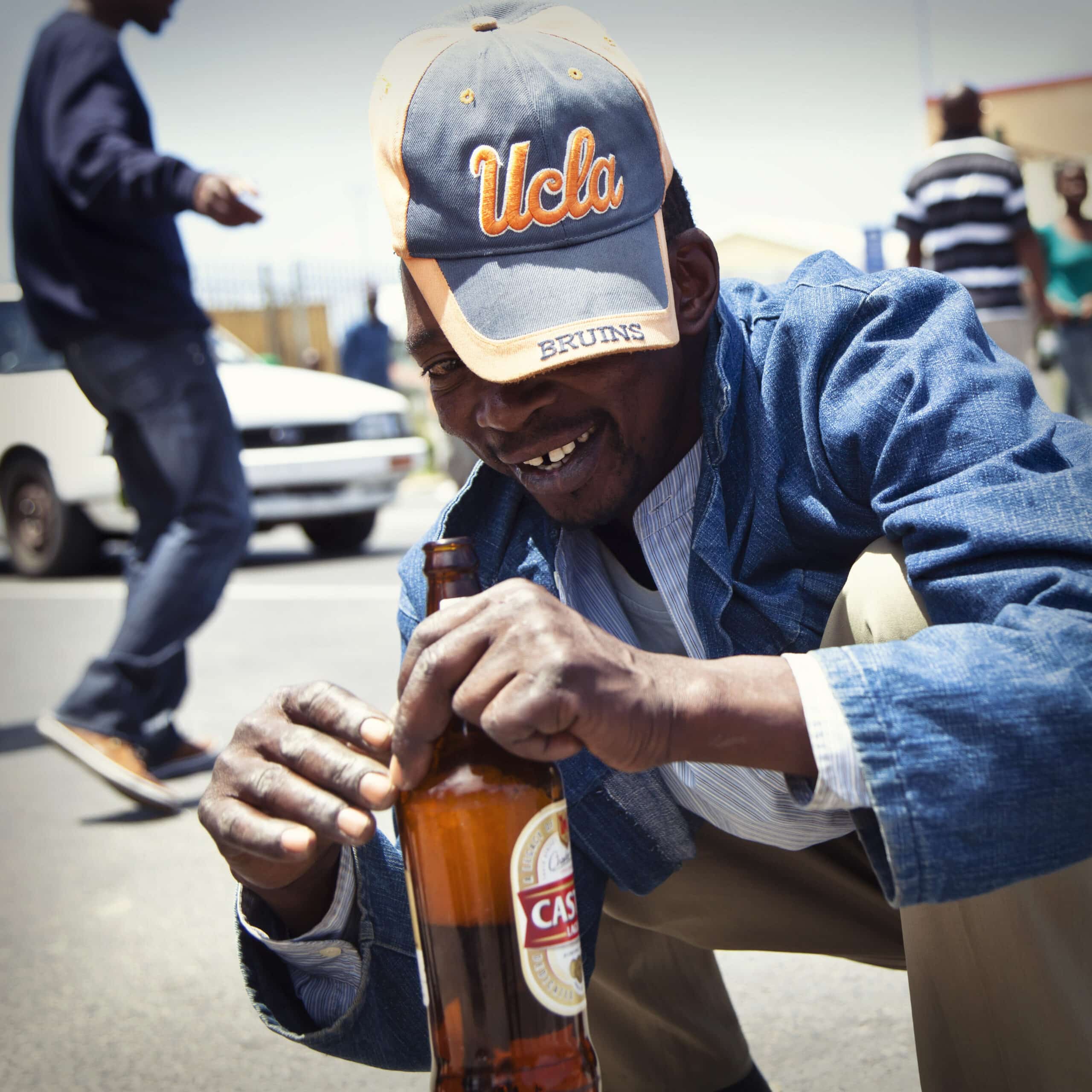 Ein Mann mit einer UCLA-Bruins-Kappe lächelt, während er auf einer sonnigen Straße eine Flasche Castle Beer öffnet, im Hintergrund ist eine weitere Person zu sehen. © Fotografie Tomas Rodriguez