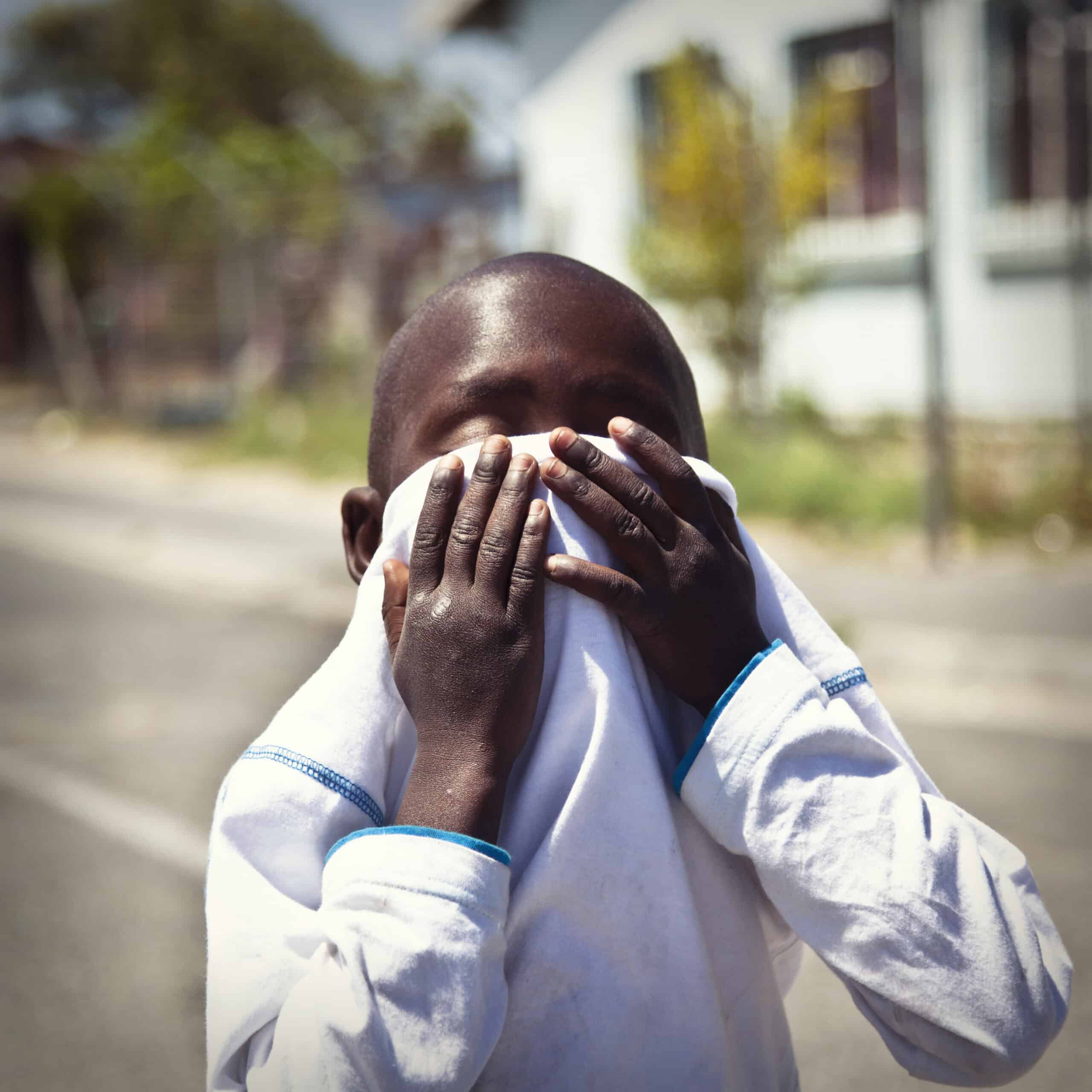 Ein kleiner Junge in einem weißen Hemd verdeckt sein Gesicht mit den Händen und steht auf einer sonnigen Straße mit verschwommenen Häusern im Hintergrund. © Fotografie Tomas Rodriguez