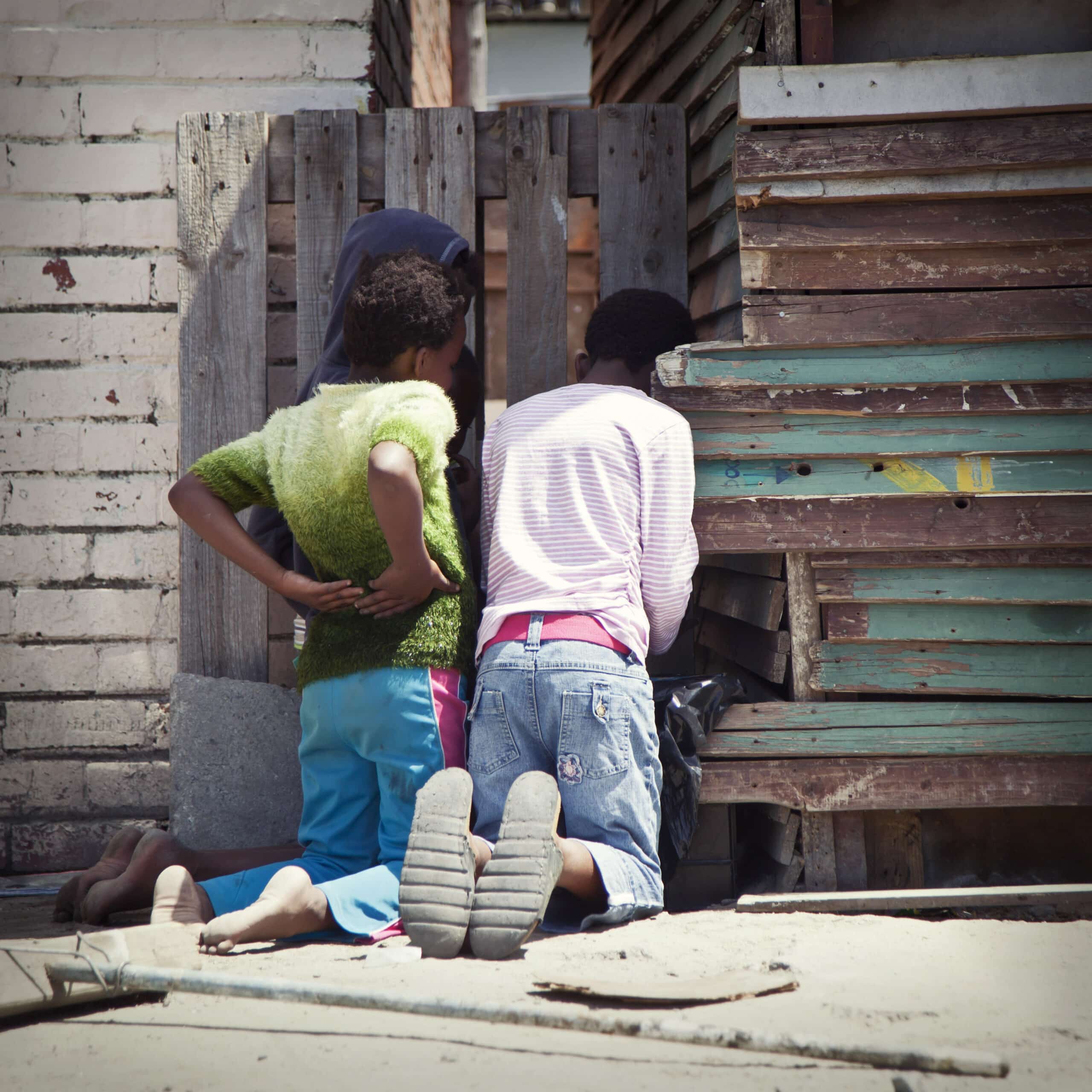 Zwei Personen sitzen vor einem improvisierten Hintergrund aus Holz zusammen. Eine trägt ein grünes Oberteil und blaue Hosen, die andere einen rosa Pullover und verwaschene Jeans. Sie scheinen in ein privates Gespräch vertieft zu sein. © Fotografie Tomas Rodriguez
