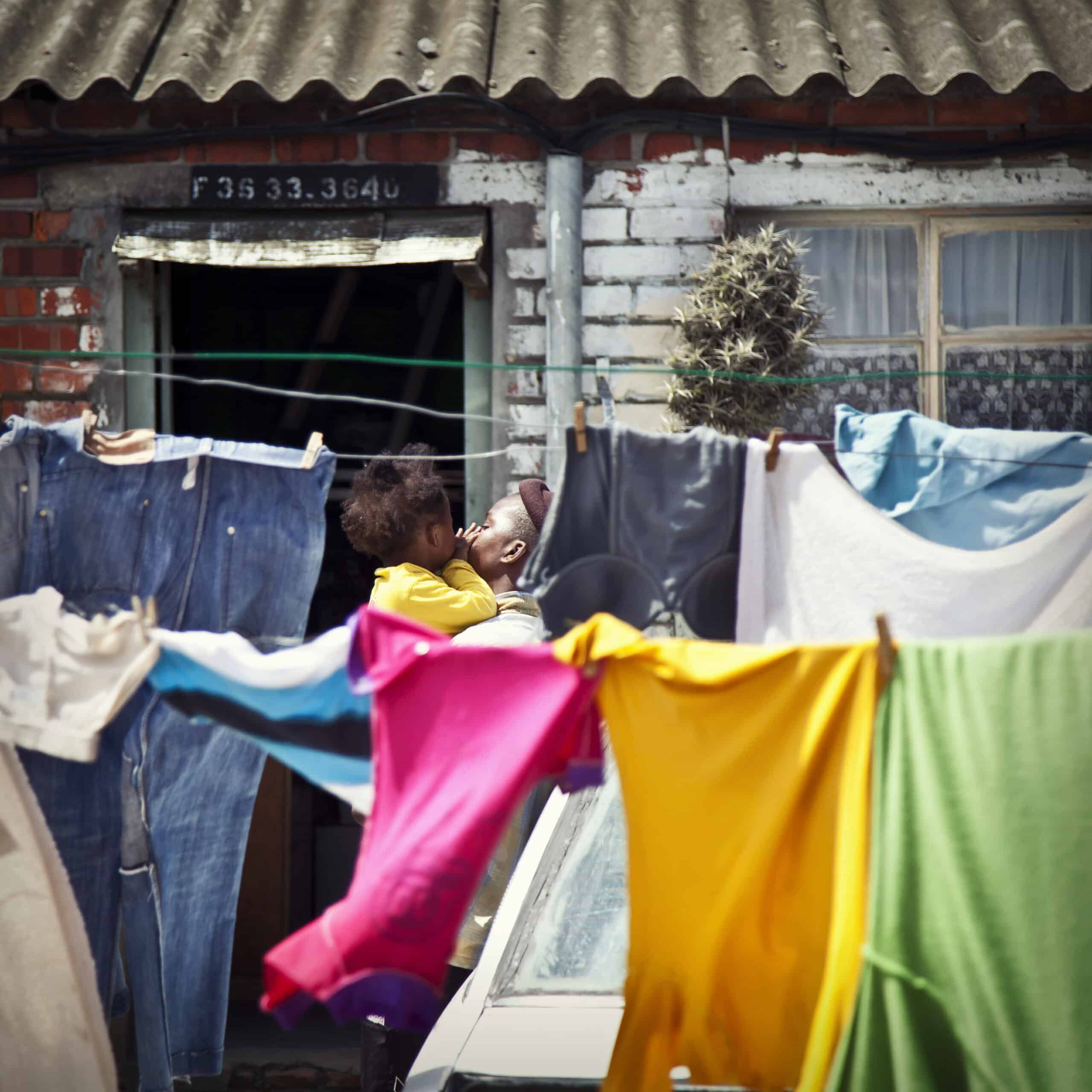 Eine Frau hält ein Kind vor einem bescheidenen Haus. Zwischen ihr und der Kamera hängt strahlend saubere Wäsche zum Trocknen auf einer Leine. © Fotografie Tomas Rodriguez
