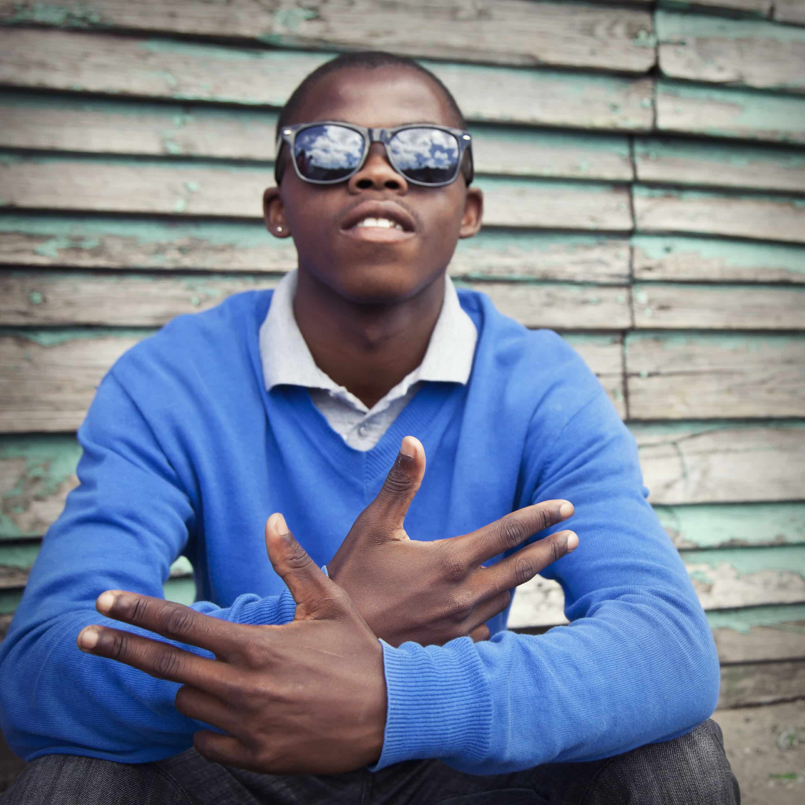 Ein junger Mann mit Sonnenbrille und blauem Pullover sitzt mit gekreuzten Fingern vor einer verwitterten Holzwand. © Fotografie Tomas Rodriguez