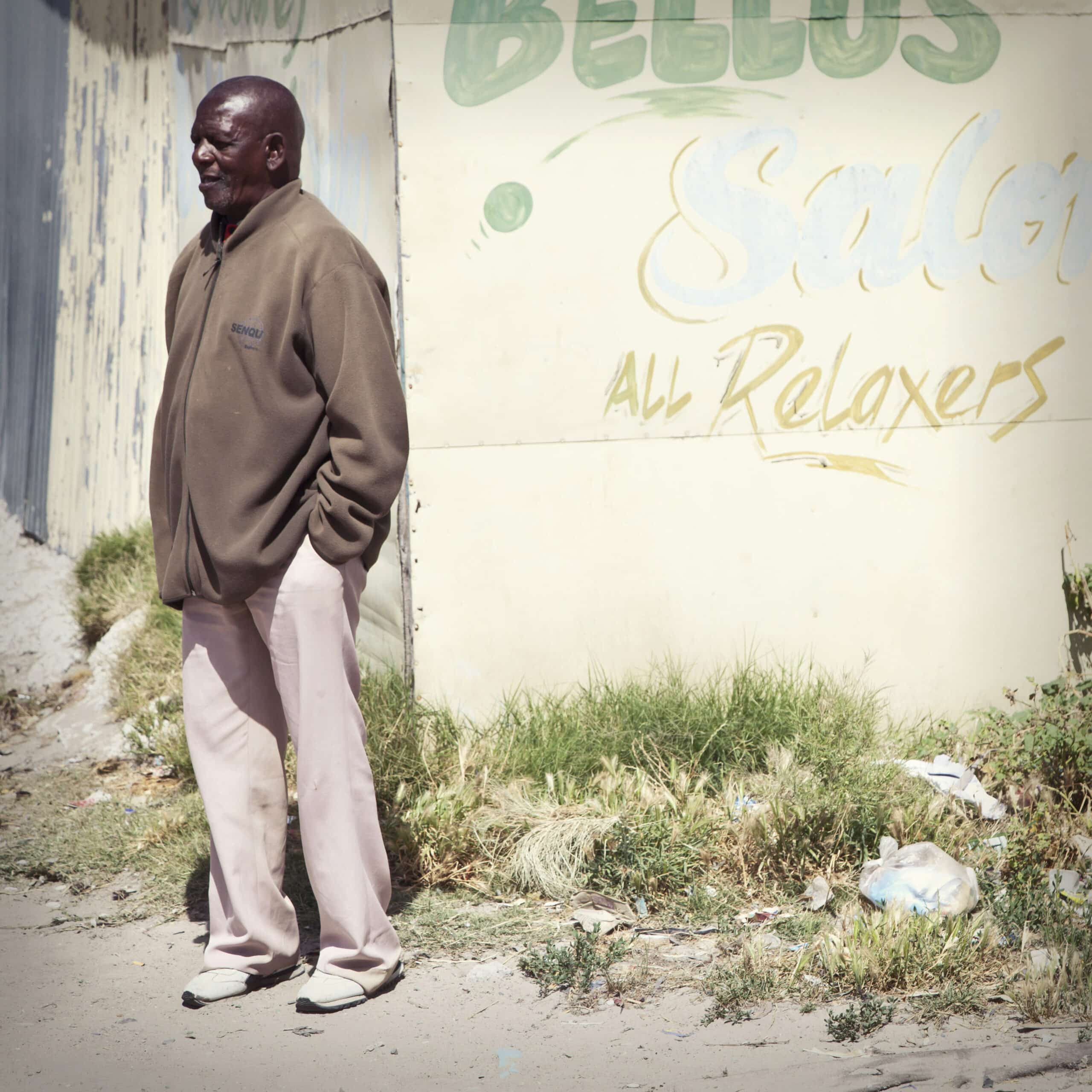 An einer verblassten Wand mit dem Graffiti „Bellus Salon & Relaxers“ steht ein älterer Mann, bekleidet mit einer braunen Jacke und hellrosa Hosen, und blickt nachdenklich zur Seite. © Fotografie Tomas Rodriguez