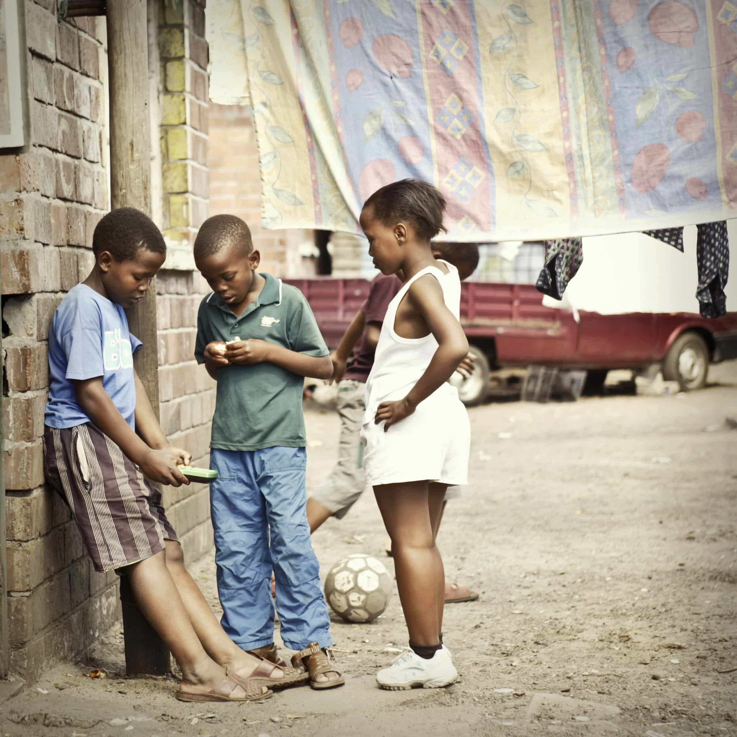 Drei Kinder in einer ländlichen Gasse, ein Mädchen und zwei Jungen, sind in Gespräche und Spiele vertieft und stehen neben einem improvisierten Fußball, in dessen Nähe bunter Stoff hängt. © Fotografie Tomas Rodriguez