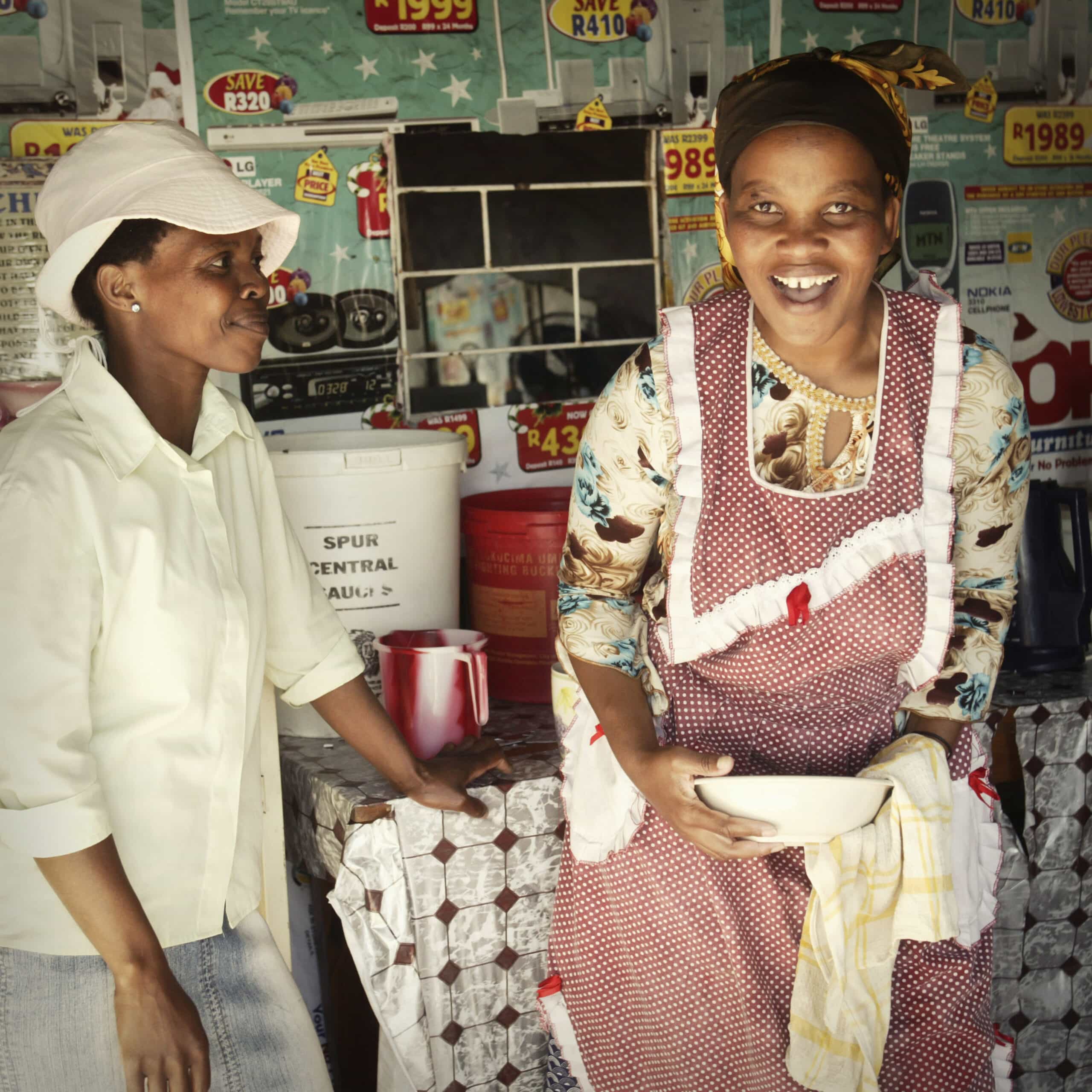 Zwei afrikanische Frauen verbringen einen freudigen Moment in einer gemütlichen, sonnendurchfluteten Küche. Die eine trägt eine Kochmütze und eine weiße Uniform und hört der anderen in einer bunten Schürze zu, die lacht und eine Schüssel hält. © Fotografie Tomas Rodriguez