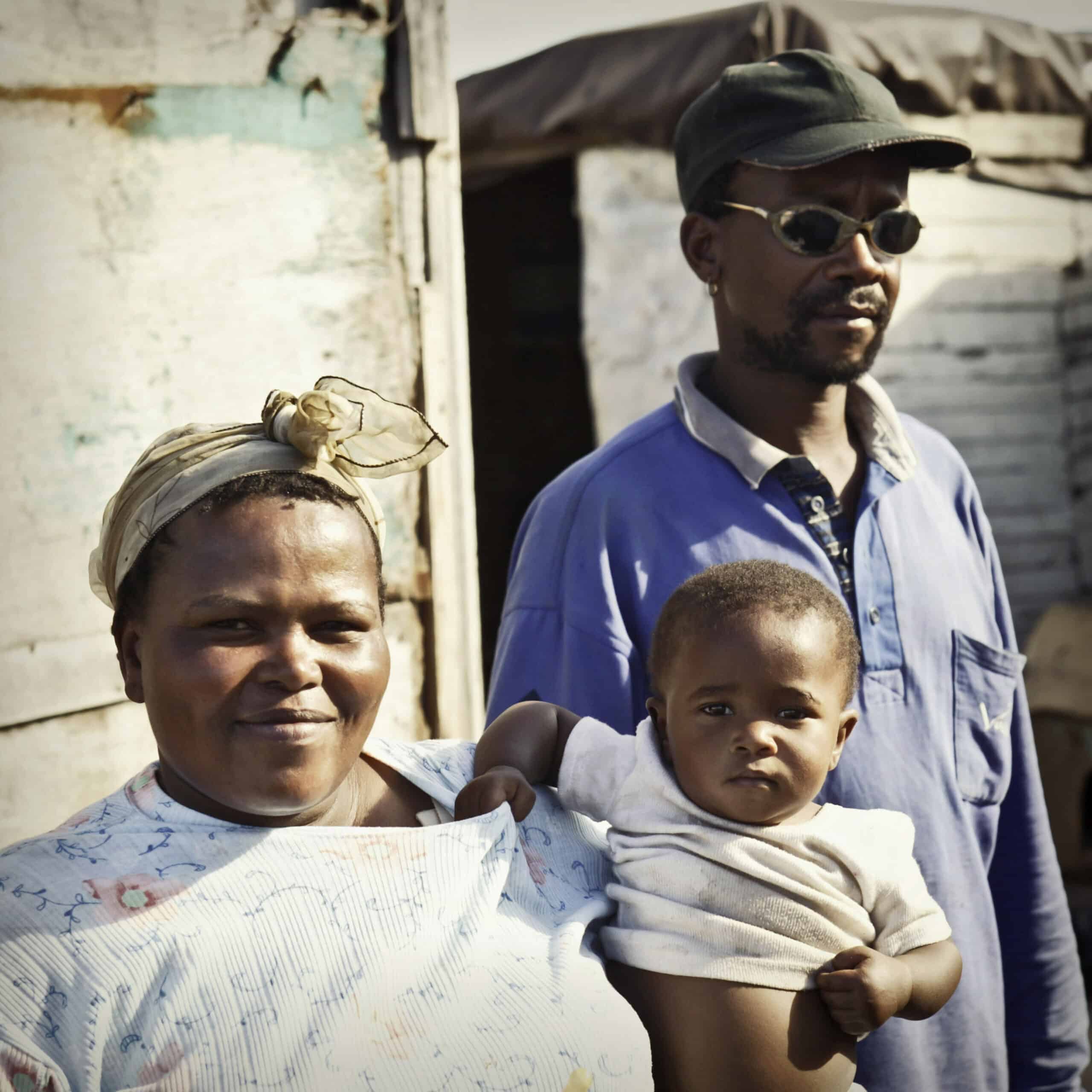 Eine Familie mit einem Mann, einer Frau und einem Baby steht vor einem Haus in einer ländlichen Umgebung. Die Frau hält das Baby und der Mann steht mit einer Sonnenbrille hinter ihnen. © Fotografie Tomas Rodriguez