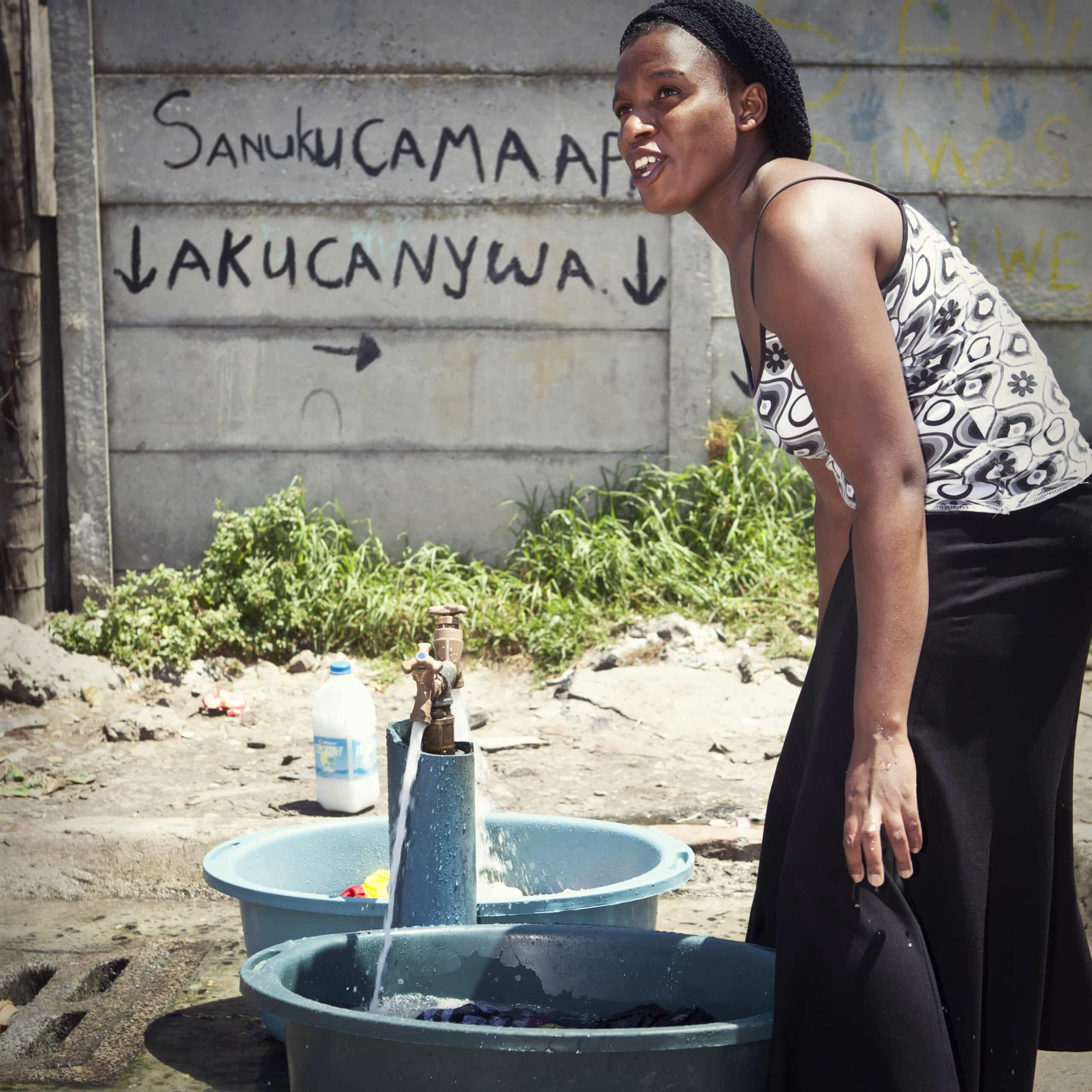 Eine Frau wäscht ihre Wäsche an einem Wasserhahn im Freien, der über mehrere mit Wasser gefüllte Becken verfügt. Ein Graffiti an der Wand hinter ihr enthält eine Botschaft in einer afrikanischen Sprache. © Fotografie Tomas Rodriguez