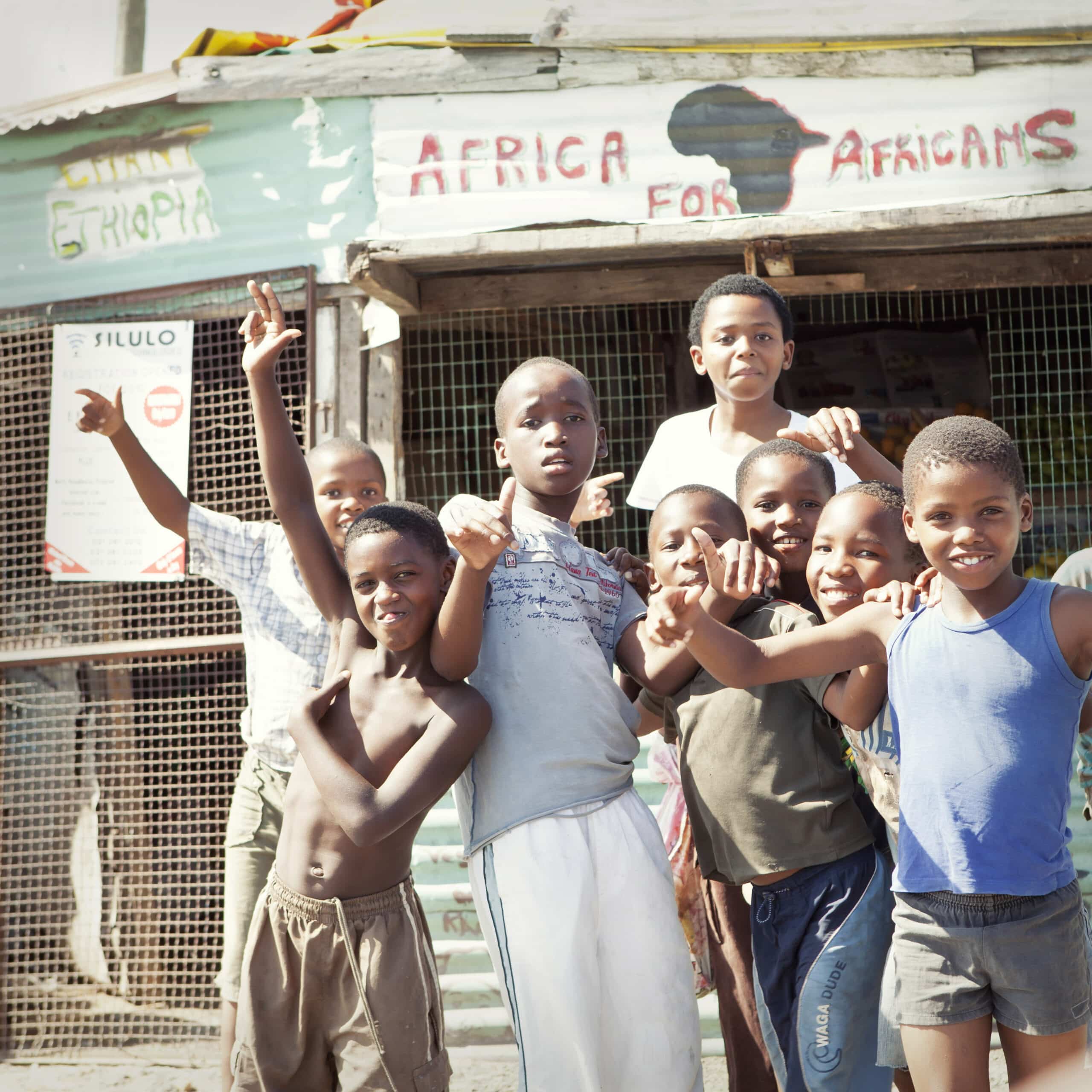 Eine Gruppe lächelnder Kinder posiert vor einem kleinen, farbenfrohen Laden namens „Afrika für Afrikaner“, wobei einige nach oben zeigen und andere verspielte Gesten machen. © Fotografie Tomas Rodriguez