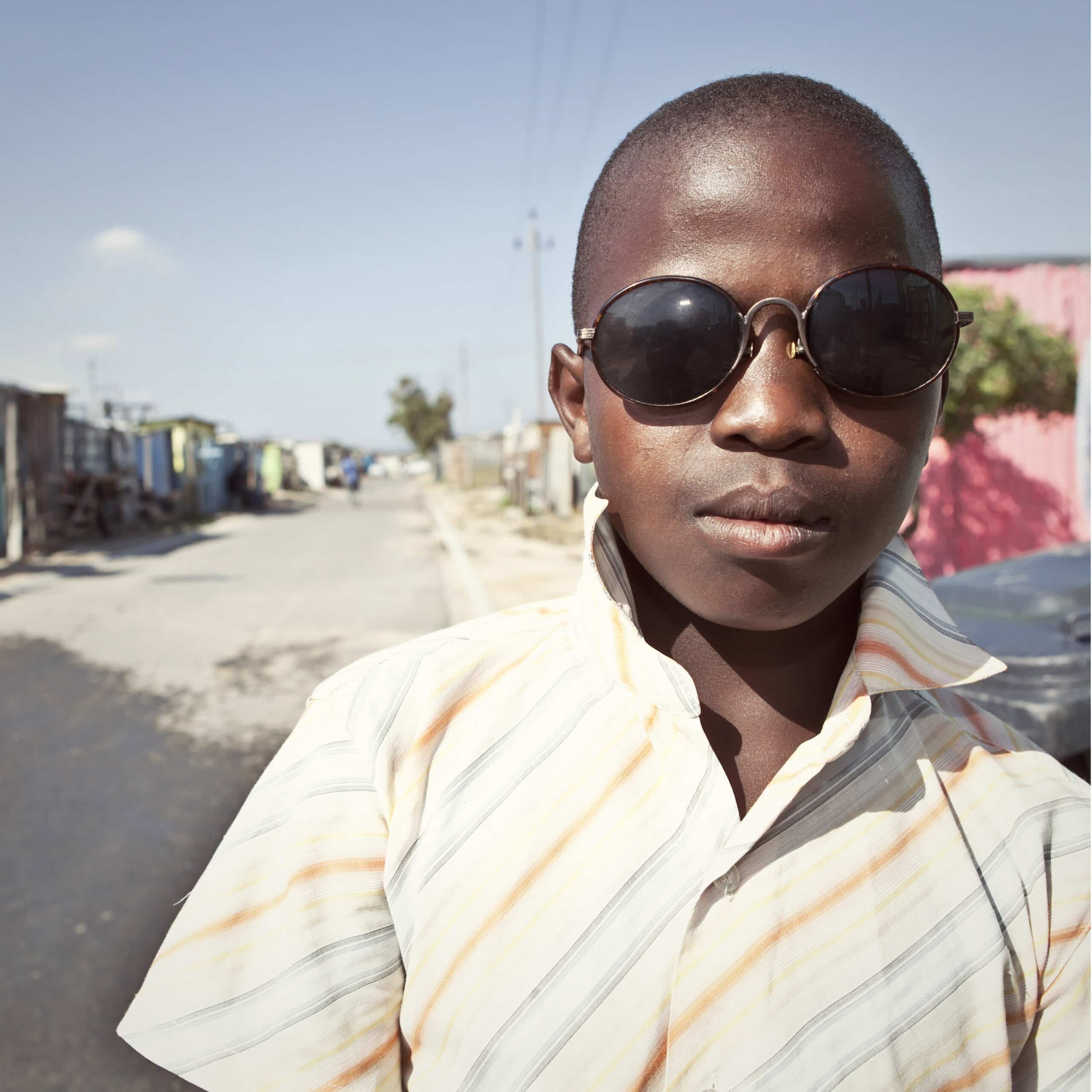 Ein kleiner Junge mit Sonnenbrille und gestreiftem Hemd steht selbstbewusst in einer sonnenbeschienenen, ungezwungenen städtischen Umgebung und blickt direkt in die Kamera. © Fotografie Tomas Rodriguez