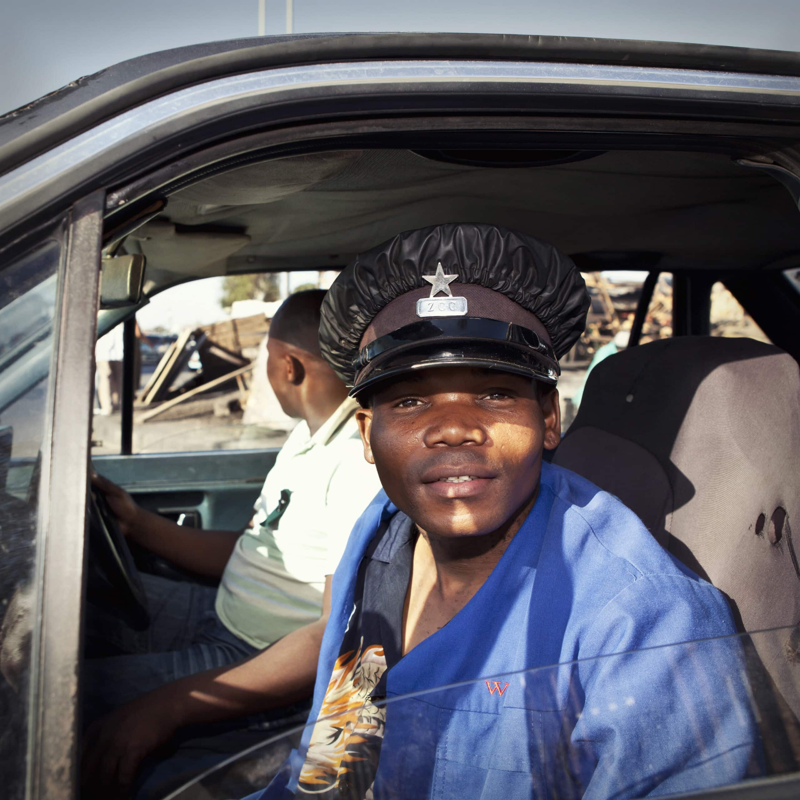 Ein Mann in einem blauen Arbeitshemd und einer schwarzen Polizeimütze sitzt auf dem Fahrersitz eines verwitterten Fahrzeugs und lächelt in einer sonnigen Umgebung in die Kamera. © Fotografie Tomas Rodriguez