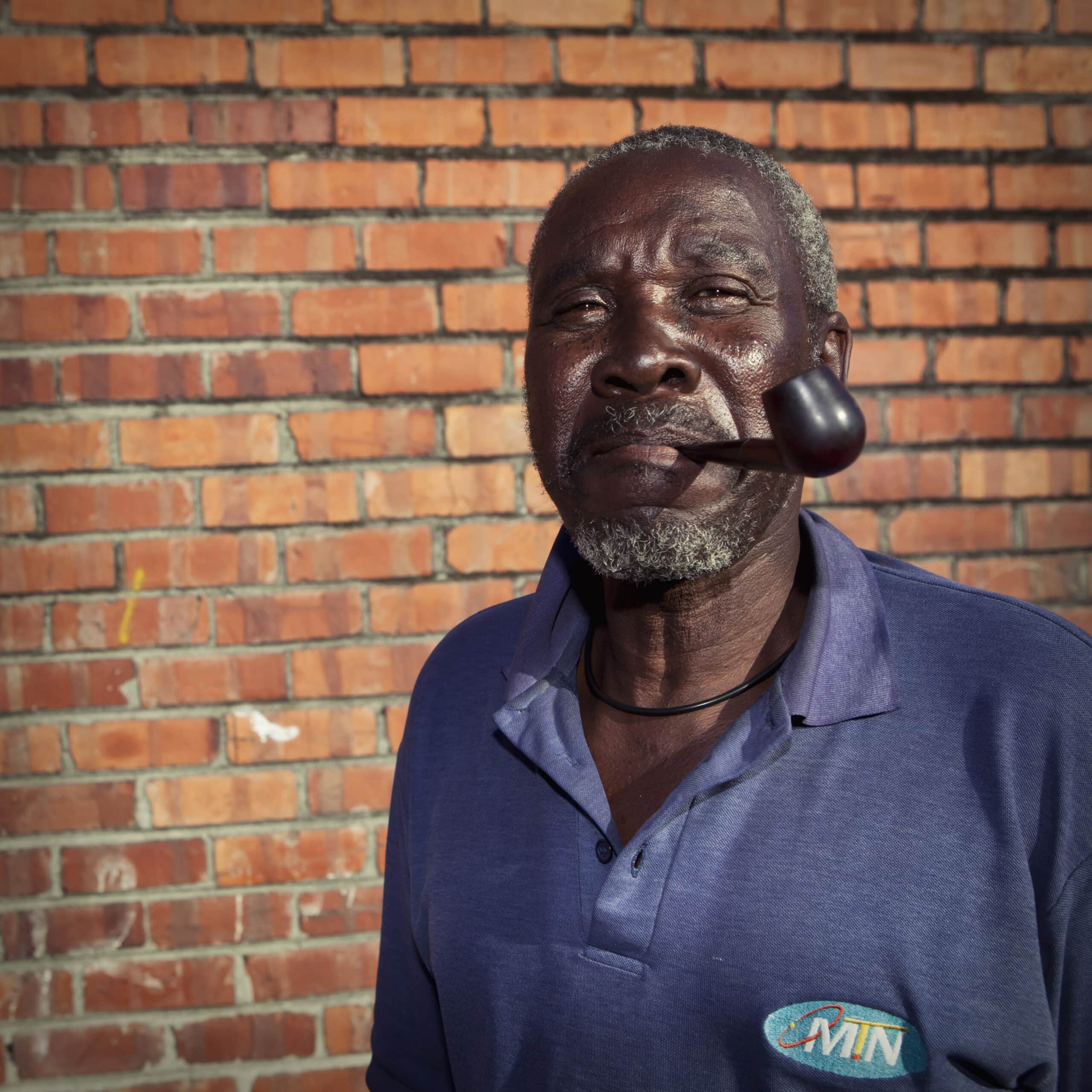 Ein älterer Mann mit verwittertem Gesichtsausdruck und grauem Haar steht vor einer Backsteinmauer und hält eine Pfeife im Mund. Er trägt ein blaues Poloshirt mit einem Logo darauf. © Fotografie Tomas Rodriguez