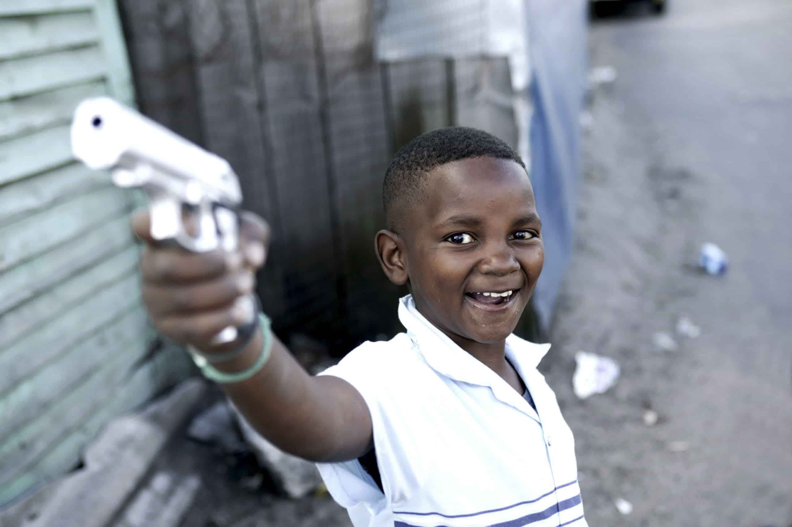 Ein kleiner Junge lächelt breit und richtet auf einer Straße eine Spielzeugpistole in die Kamera. Er trägt ein hellblaues Hemd. Der verschwommene Hintergrund suggeriert eine städtische Umgebung. © Fotografie Tomas Rodriguez