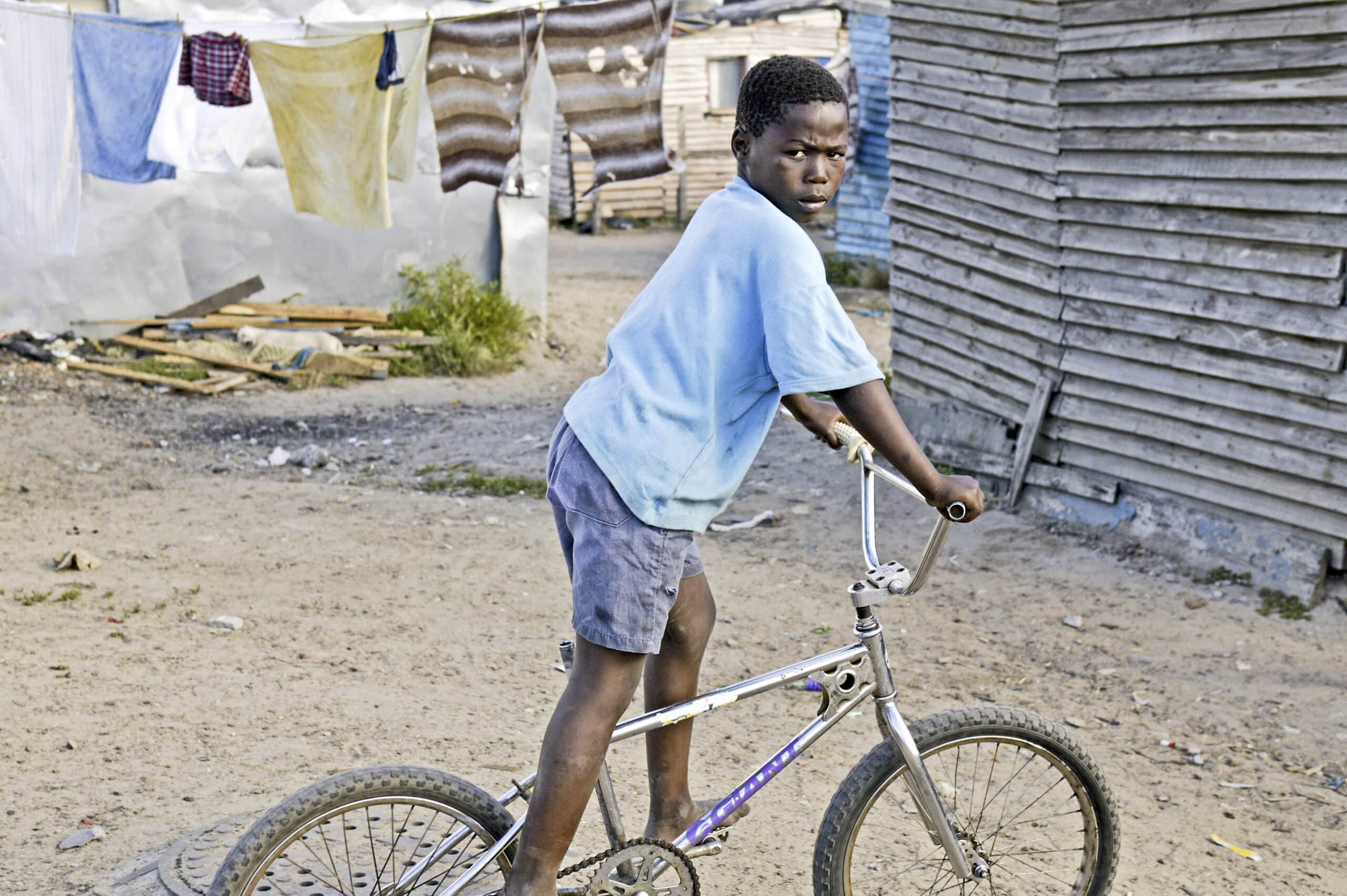 Ein kleiner Junge steht mit seinem Fahrrad in einer Elendsviertelgegend. Im Hintergrund hängen Kleider und überall liegt Schutt herum. Er blickt mit ernster Miene zurück. © Fotografie Tomas Rodriguez