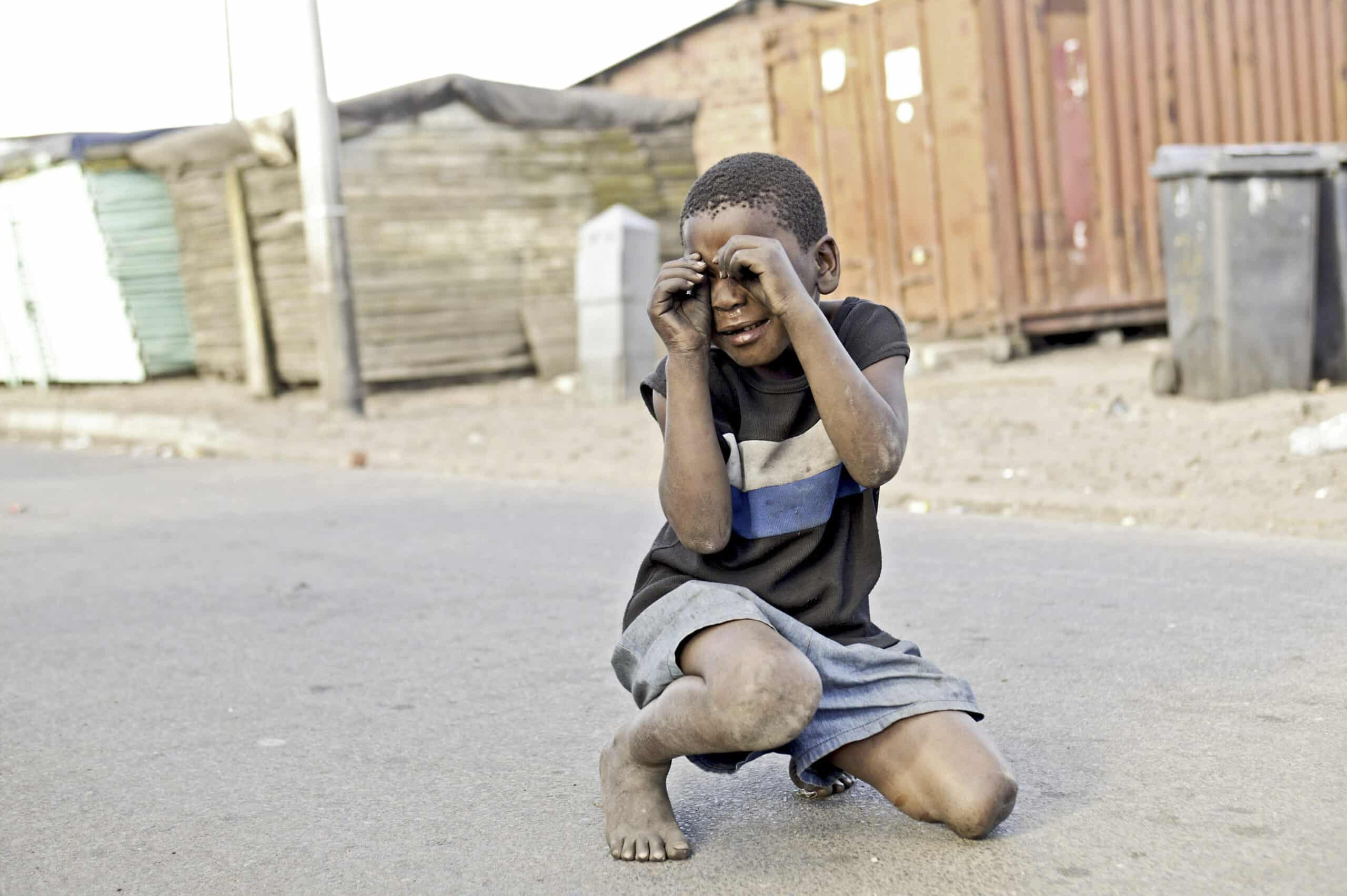 Ein kleiner Junge hockt auf einer staubigen Straße, formt mit seinen Händen vor den Augen Fingergläser und lächelt spielerisch. Im Hintergrund sind provisorische Behausungen und Schiffscontainer zu sehen. © Fotografie Tomas Rodriguez