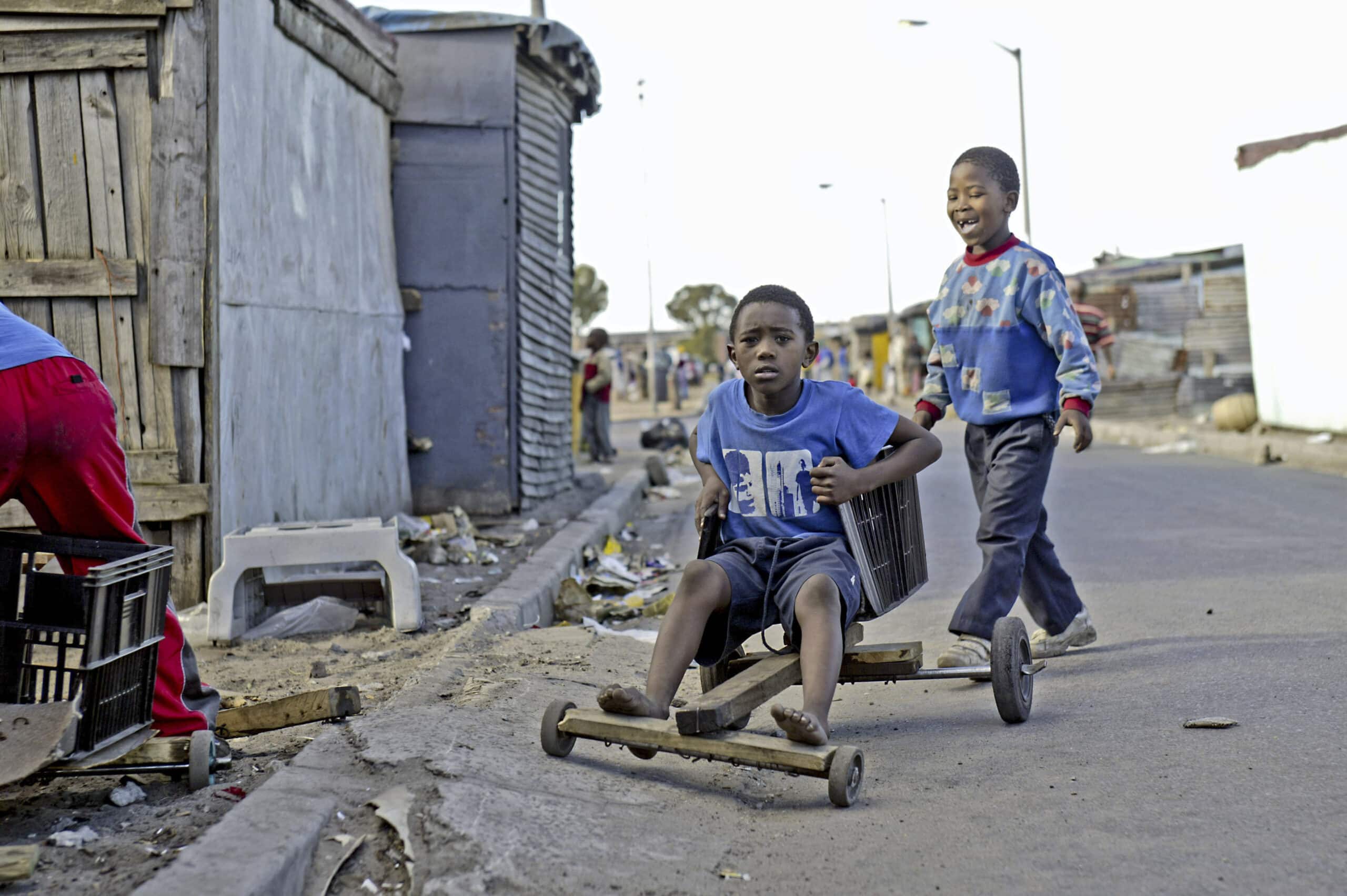 Zwei Jungen spielen mit einem selbstgebauten Go-Kart aus Holz auf einer staubigen Straße. Ein Junge schiebt das Kart an, ein anderer sitzt darin und lenkt es mit einer Schnur. Vorstädtische Kulisse mit informellen Strukturen. © Fotografie Tomas Rodriguez
