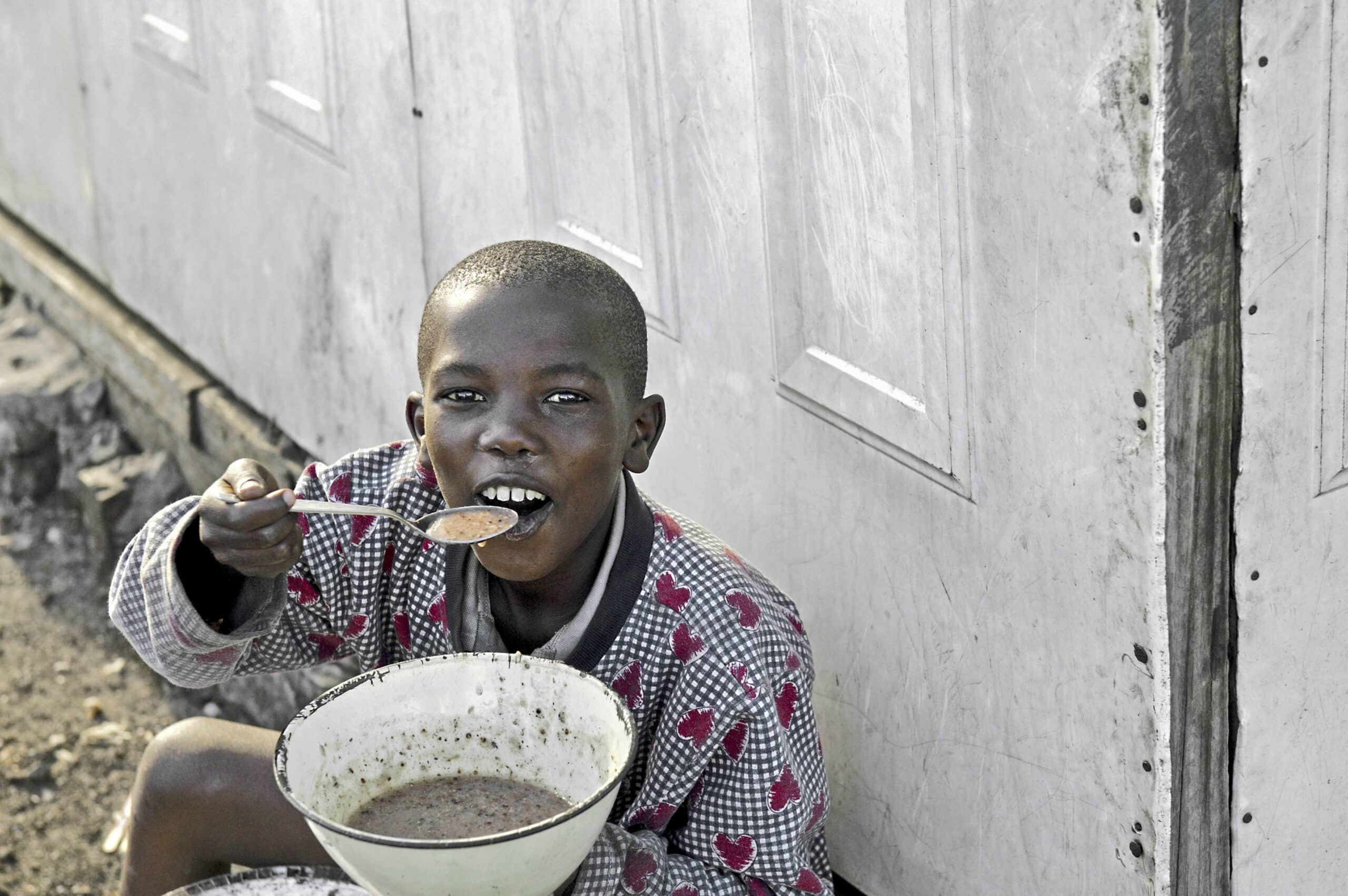 Ein kleiner Junge in einem rot gemusterten Hemd isst mit einem Löffel aus einer Schüssel und sitzt im Freien an einer grauen Wand. Sein Gesichtsausdruck ist zufrieden und er konzentriert sich auf sein Essen. © Fotografie Tomas Rodriguez