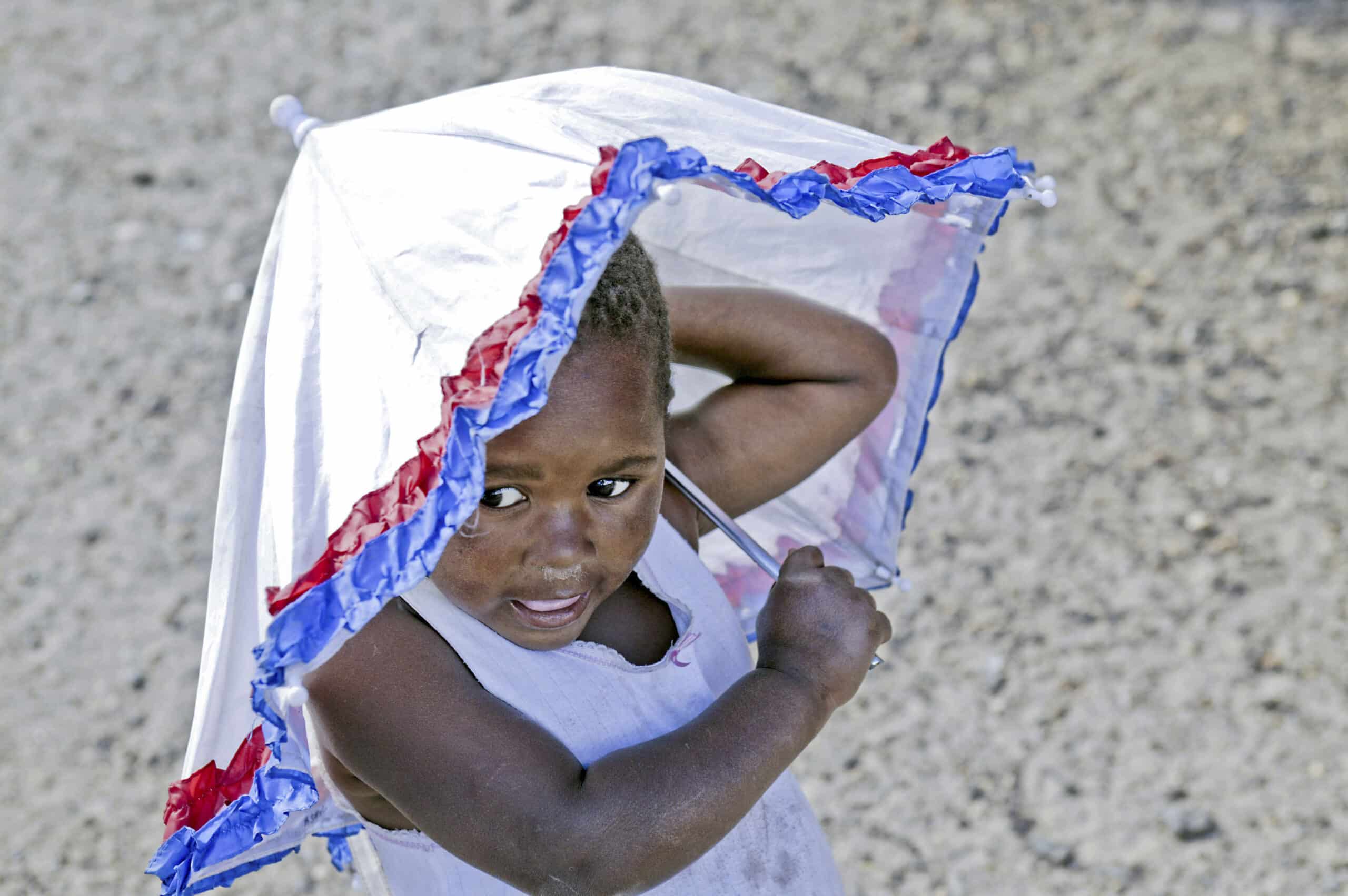 Ein kleines Kind steht vor einem Kieshintergrund und hält einen weißen Drachen mit blauen und roten Rändern über dem Kopf, während es mit neugierigem Gesichtsausdruck nach oben blickt. © Fotografie Tomas Rodriguez