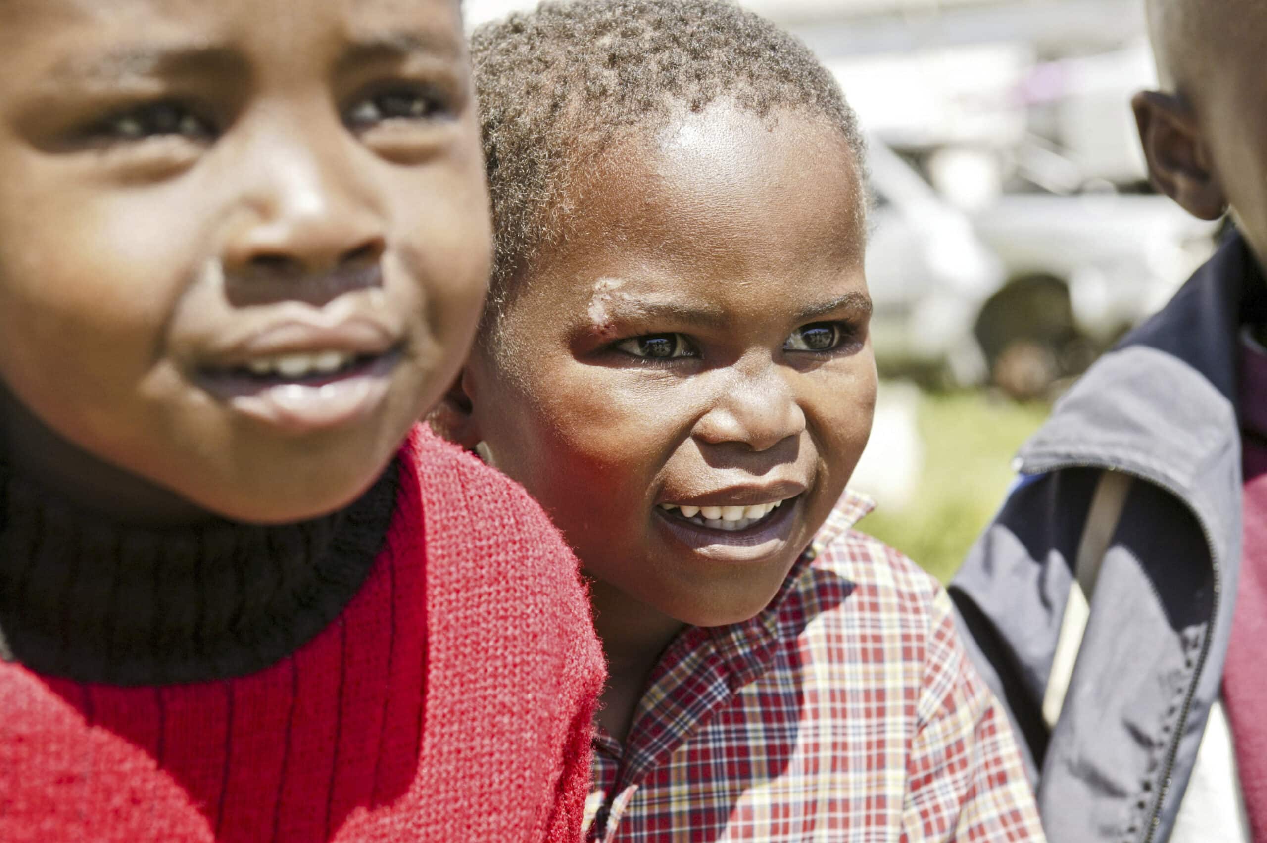 Zwei kleine Kinder im Freien, im Mittelpunkt ein lächelndes Kind im roten Pullover und ein anderes im karierten Hemd. Helles Sonnenlicht verstärkt ihre freudigen Ausdrücke. © Fotografie Tomas Rodriguez