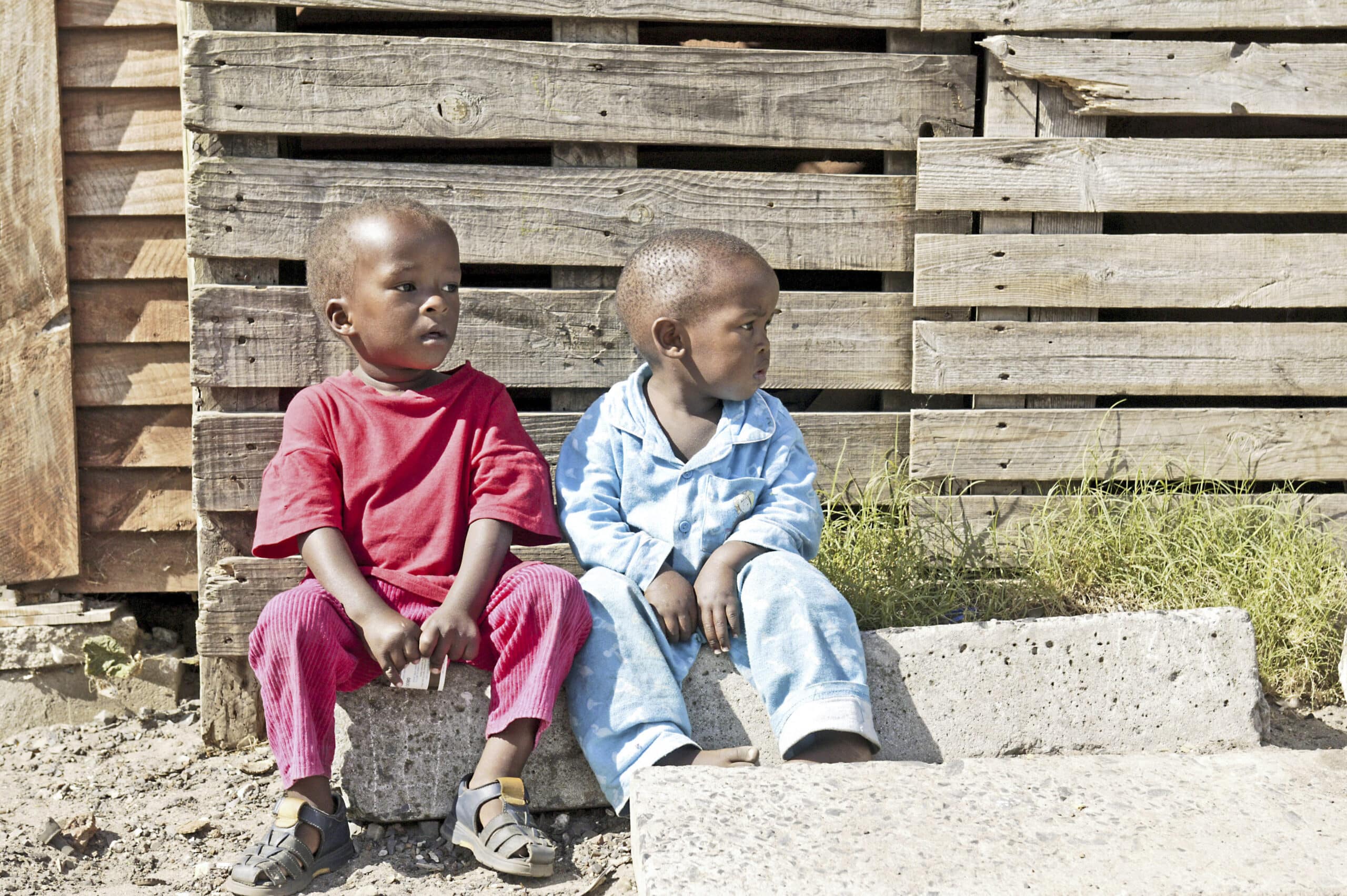 Zwei kleine Kinder, eines im roten Hemd und eines im blauen Outfit, sitzen nebeneinander auf einem Betonblock neben einer Wand aus Holzpaletten in einer sonnigen Umgebung im Freien. © Fotografie Tomas Rodriguez