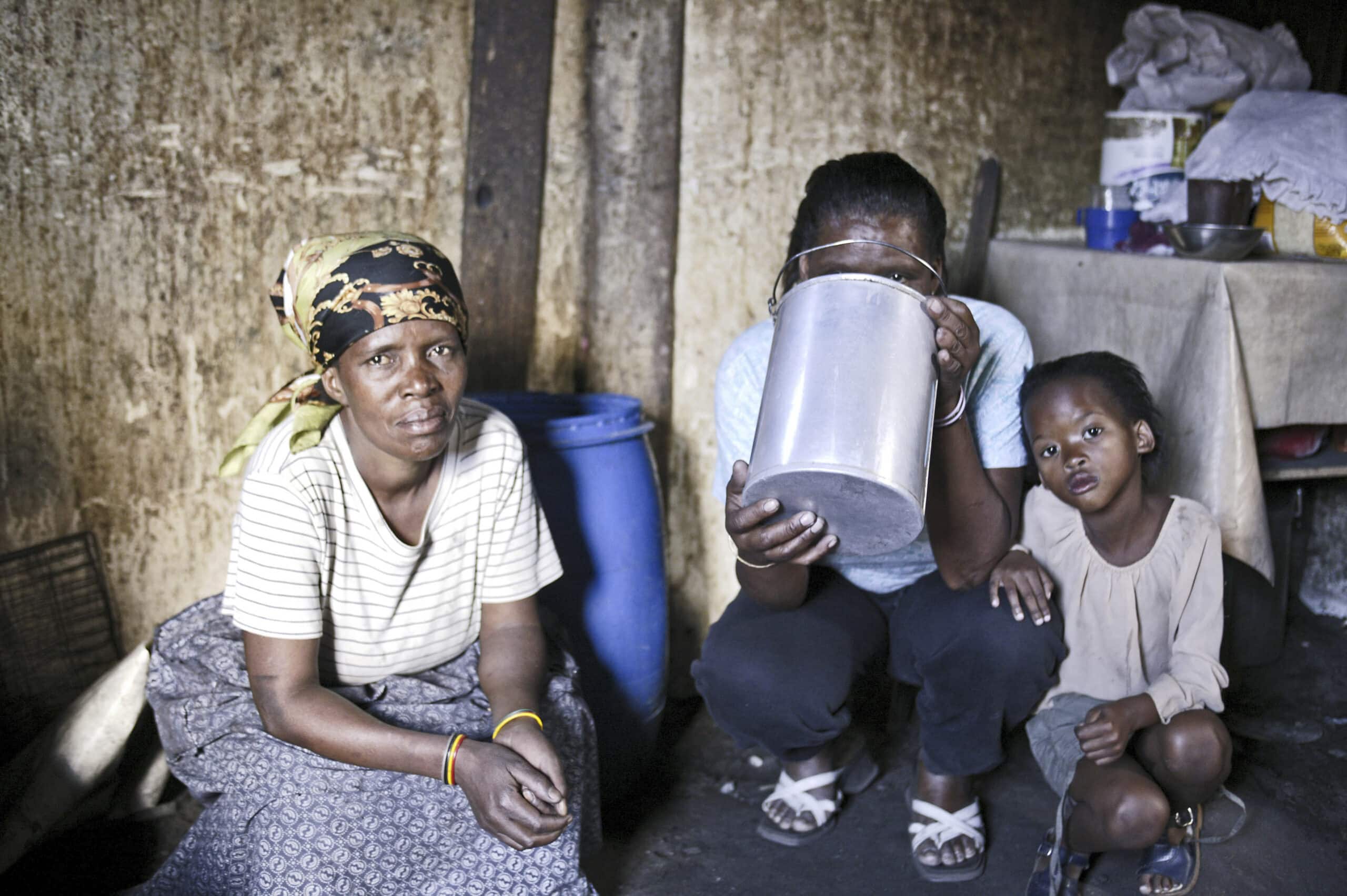 Eine Familie in einem schwach beleuchteten Raum. Eine Frau sitzt da und hält ein kleines Kind, neben ihnen steht ein Mann, der Wasser aus einem Krug gießt. © Fotografie Tomas Rodriguez