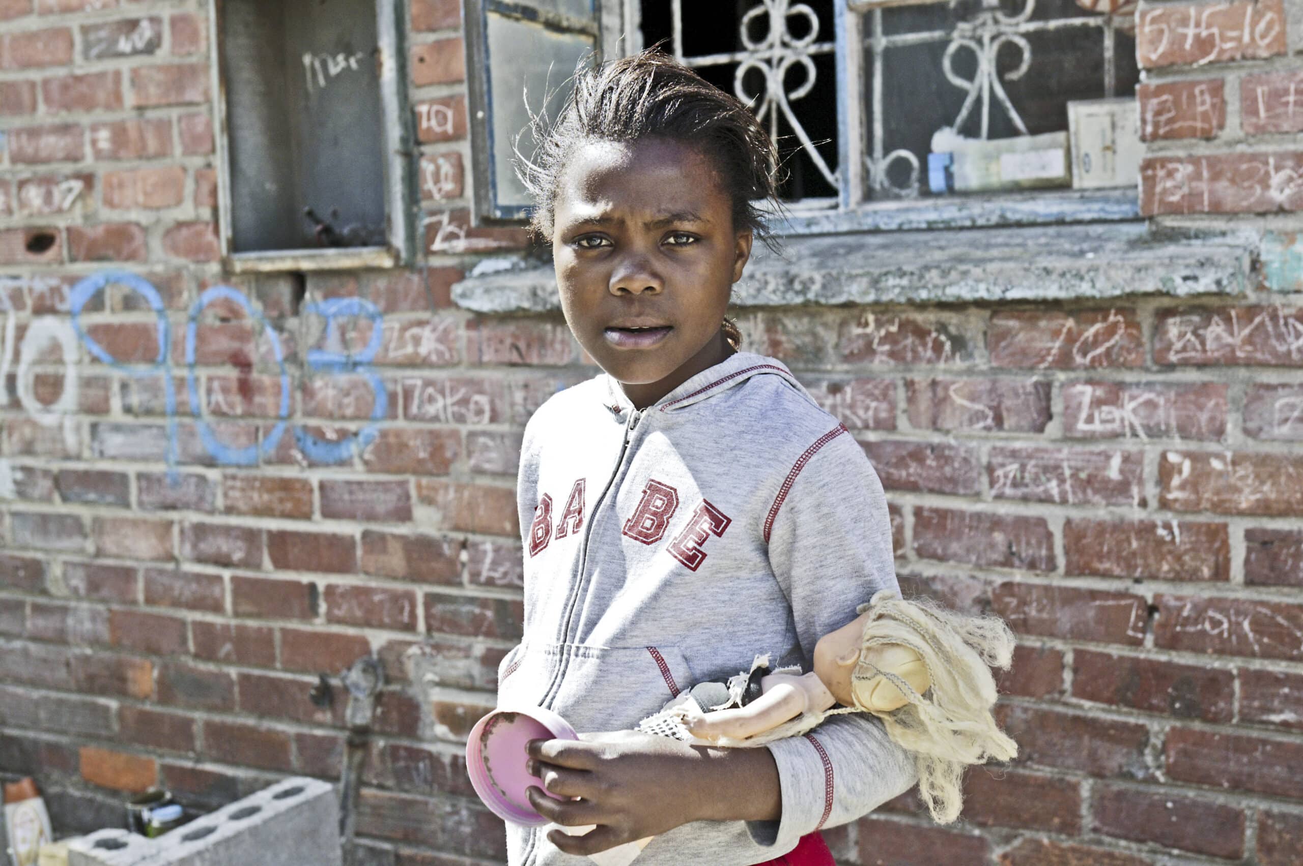 Ein junges Mädchen mit ernster Miene steht vor einer Backsteinmauer mit Graffiti, hält eine Puppe und einen rosa Gegenstand und trägt einen grauen Kapuzenpullover mit der Aufschrift „Babe“. © Fotografie Tomas Rodriguez