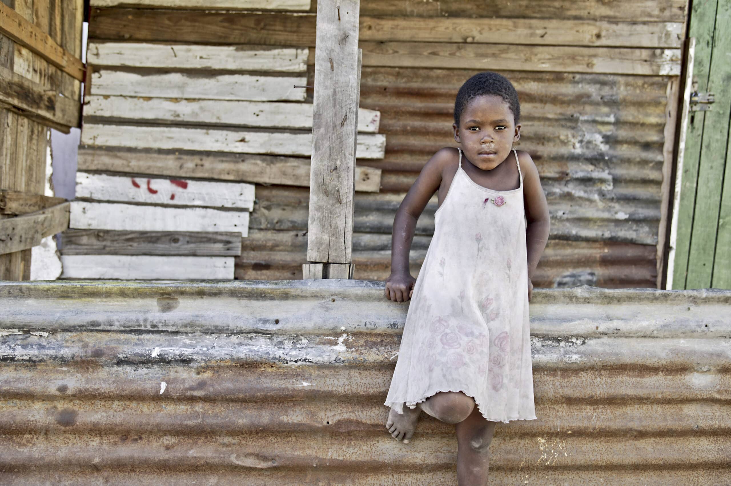 Ein kleines Kind in einem verblichenen Kleid steht barfuß und lehnt an einer verwitterten Metallbarriere vor einer Holzhütte. © Fotografie Tomas Rodriguez