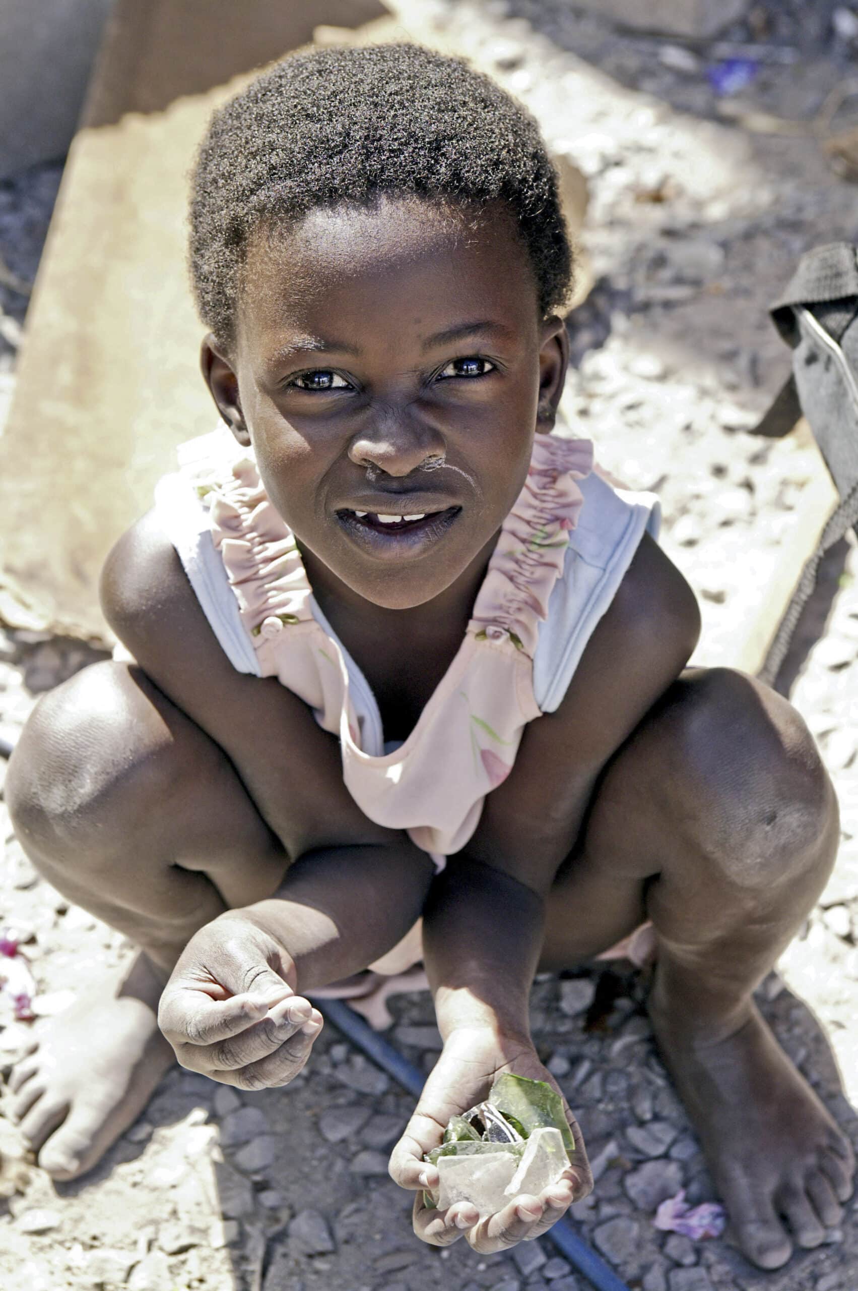 Ein kleines Kind mit kurzen Haaren und einem hellrosa Kleid hockt auf dem Boden und hält einen kleinen Gegenstand. Der Hintergrund zeigt einen sandigen und steinigen Untergrund. © Fotografie Tomas Rodriguez
